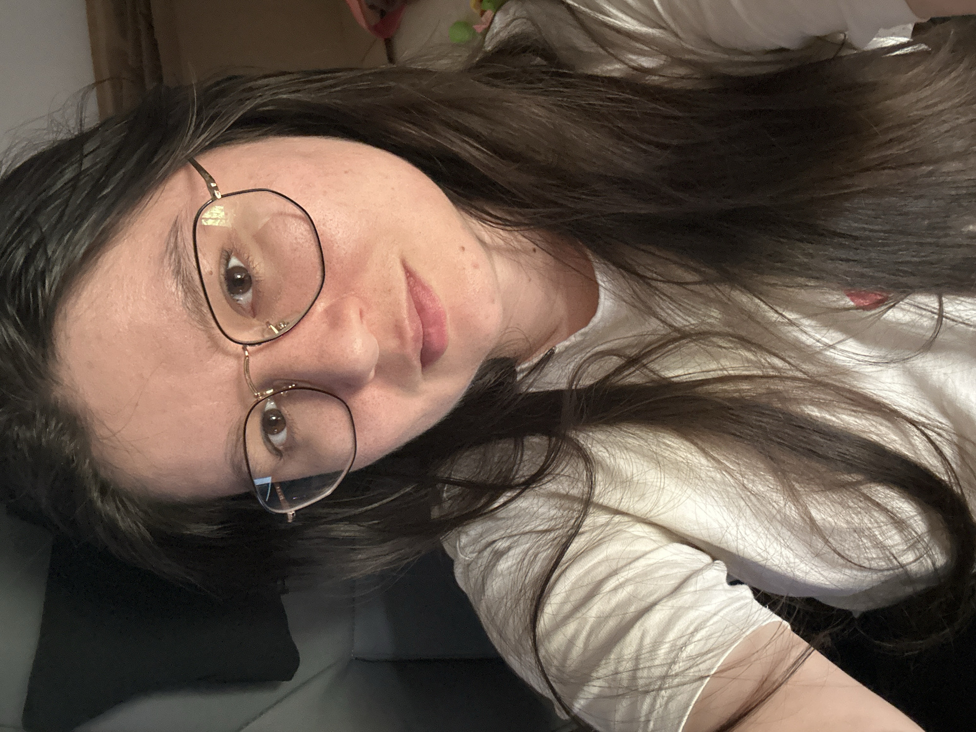 A woman with long, dark brown hair and wearing glasses is shown in a close-up selfie. She is lying down with her head tilted, looking directly at the camera. She has fair skin and is wearing a white t-shirt. The background is blurry, but appears to be a black surface. No indication of the location being Câmara de Lobos, Portugal is present in the image.