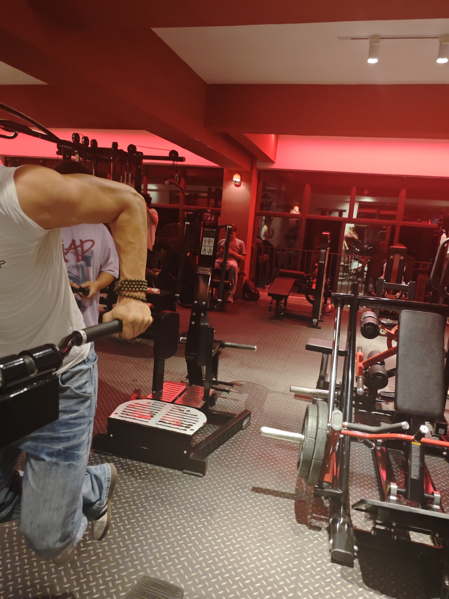 The image captures a gym interior in Chongqing High-tech Zone, China, characterized by a red lighting scheme. Several individuals are present; one is actively using a seated cable row machine, wearing a white tank top and jeans, and adorned with beaded bracelets. Another person, wearing a t-shirt with a graphic print, is observing nearby.
Gym equipment is prominent, including the seated cable row machine in use, and a leg extension/curl machine. Other machines, such as a rowing machine and weight benches, are visible in the background through a glass partition. The gym floor has a diamond plate pattern.