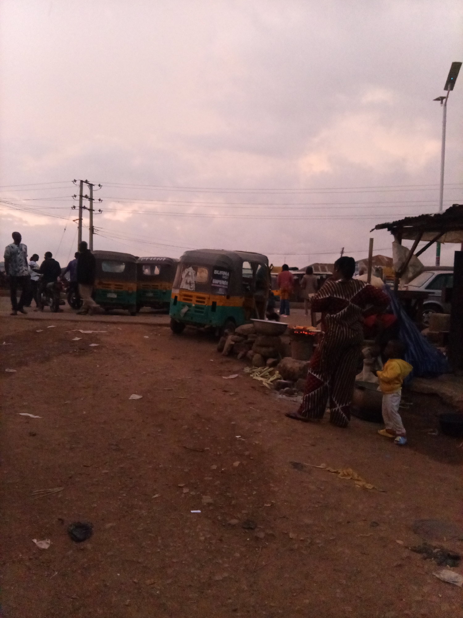 An eye-level, outdoor, medium shot captures a street scene with several people and vehicles under a cloudy sky at dusk. In the foreground, the ground is a mix of reddish-brown dirt and scattered debris. To the right, a person in a striped brown and white outfit stands near a small fire pit where food is being cooked. A small child in a yellow top and white pants stands beside them. Behind this group, a rustic stall with a corrugated metal roof is visible. Further back, a green and yellow auto-rickshaw is parked on the left, and another similar vehicle is positioned behind it. A group of people are gathered around a motorcycle on the far left. Power lines and poles are visible in the background, along with a street lamp on the right. The overall lighting suggests the image was taken at dawn or dusk.