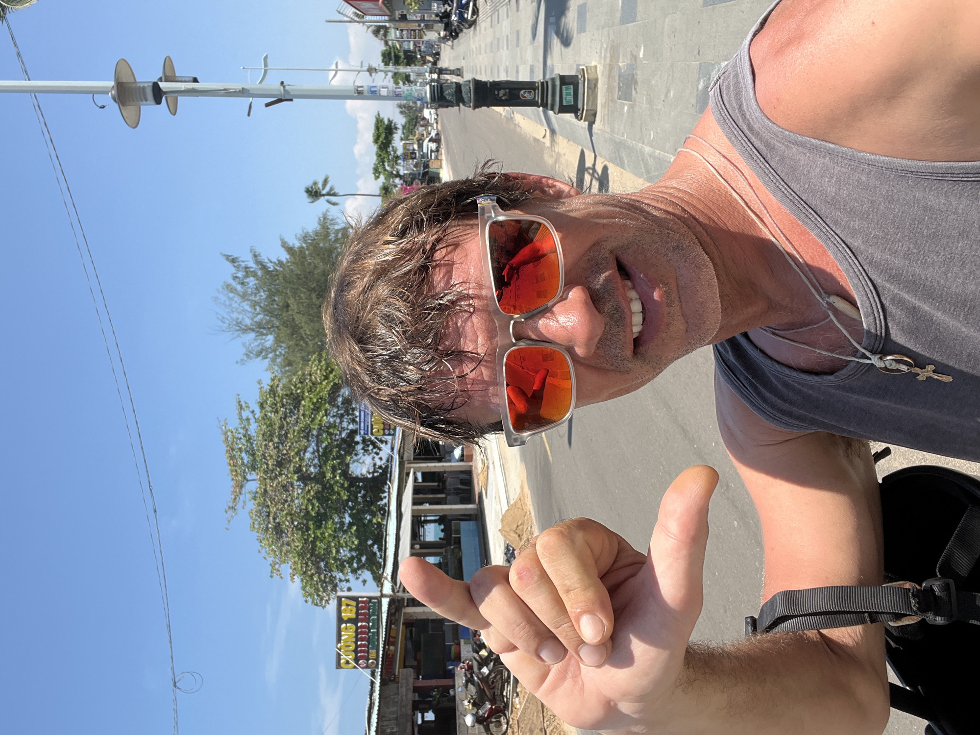 A man is taking a selfie on a sunny day. He is wearing a gray tank top and sunglasses with red lenses. His right hand is making a shaka sign. In the background, there is a street with buildings and palm trees. The man appears to be in Phu Thuy Ward, Vietnam, indicated by the Vietnamese text on the signs in the background.
