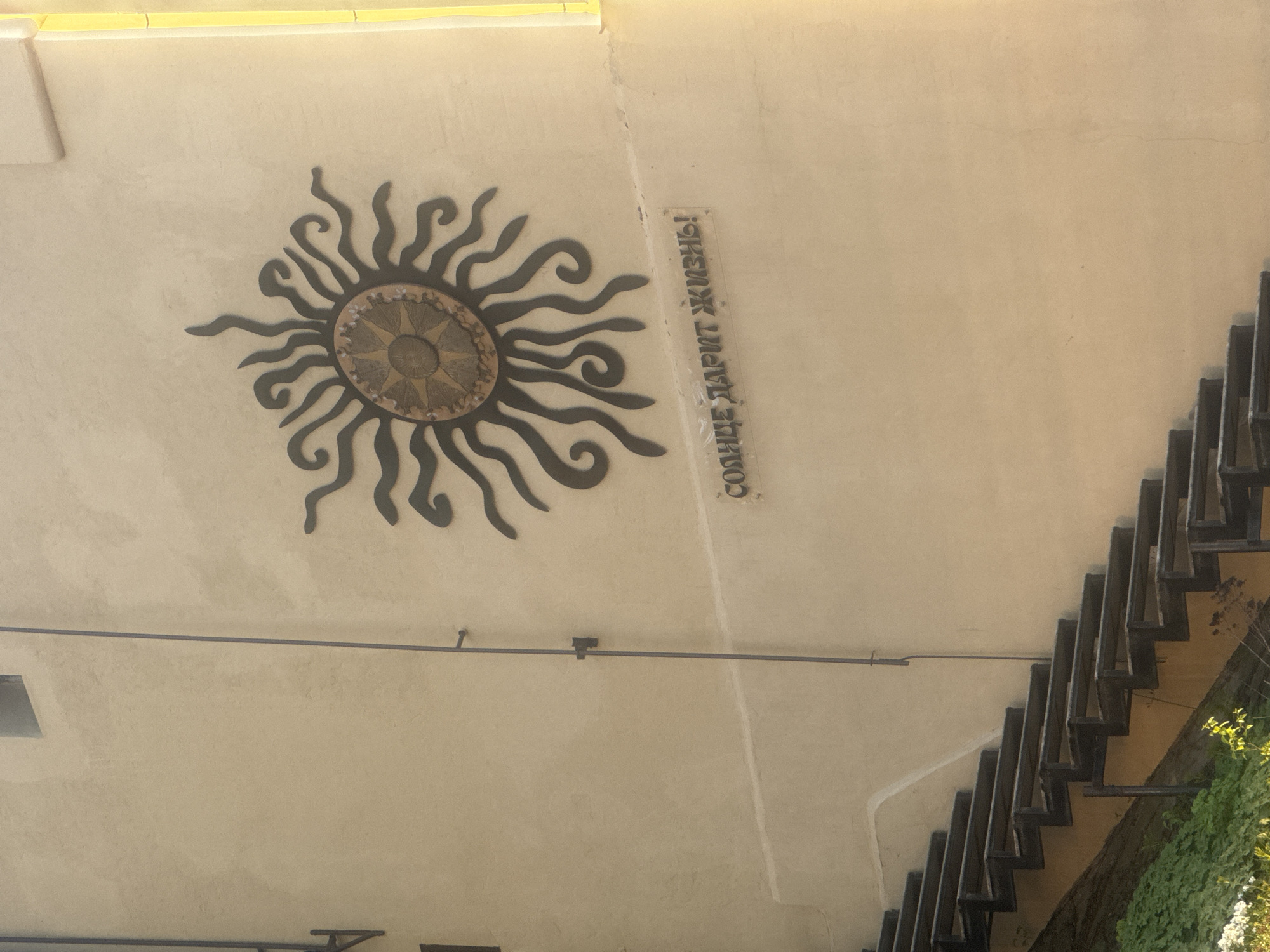 Here's a description of the image:
The image shows a section of a building exterior in Nizhny Novgorod, Russia. A decorative sun motif, likely made of metal, is affixed to a light-colored wall. To the right of the sun is a vertical inscription in Russian, "Солнце дарит жизнь!" (The sun gives life!). Below the sun and inscription is a window and what appears to be a support beam. To the right of the image are some stairs attached to the building. Some greenery is visible beside the stairs.