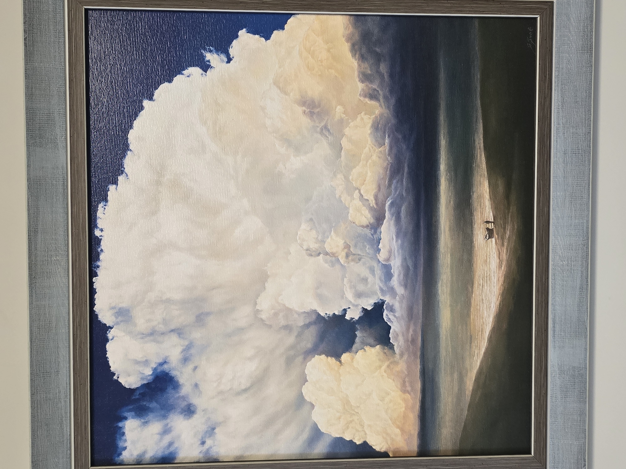 A framed painting depicting a vast cloud formation dominates the left side of the artwork, contrasted by a vertical band of land or beach on the right. The sky behind the clouds is a deep blue, transitioning to lighter hues within the clouds themselves. The land is rendered in muted tones, with two figures standing near the center-right edge. The frame around the painting is a double frame. The location is not discernible from the image.