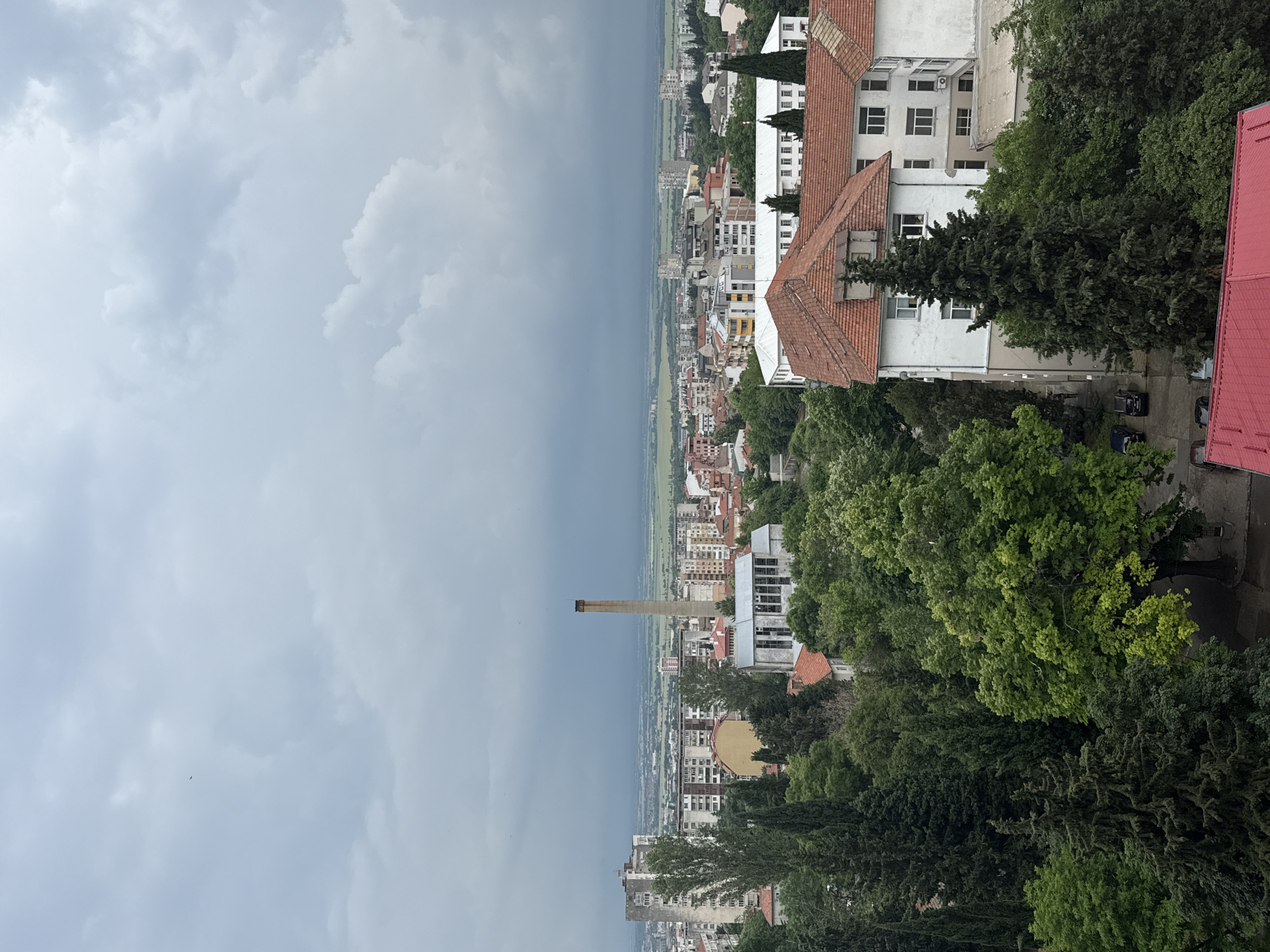This media file offers a high-angle panoramic view of a city, consistent with Stara Zagora, Bulgaria, looking out towards a distant plain under an overcast sky. The foreground and right side are dominated by lush, dense green trees and foliage, suggesting the viewpoint is from a higher elevation overlooking the urban area.
The cityscape unfolds with a mix of buildings, including multi-story structures with white facades and distinctive red-tiled roofs, alongside more modern, block-like apartment buildings. A prominent, tall, slender brick chimney or smokestack stands out in the middle distance, a solitary vertical element against the horizon. Beyond the immediate urban sprawl, the land flattens into a vast, open plain that stretches into the distance.
The time of day appears to be daytime, though the heavy, dark grey clouds create a diffused, somewhat somber light, suggesting overcast weather or an approaching storm. No direct sunlight is visible, and shadows are soft or non-existent. No people are visible in the scene, and no specific activities are taking place; it is a static landscape shot. Several cars can be faintly observed parked amongst the trees near the buildings in the foreground. There is no visible text within the image.