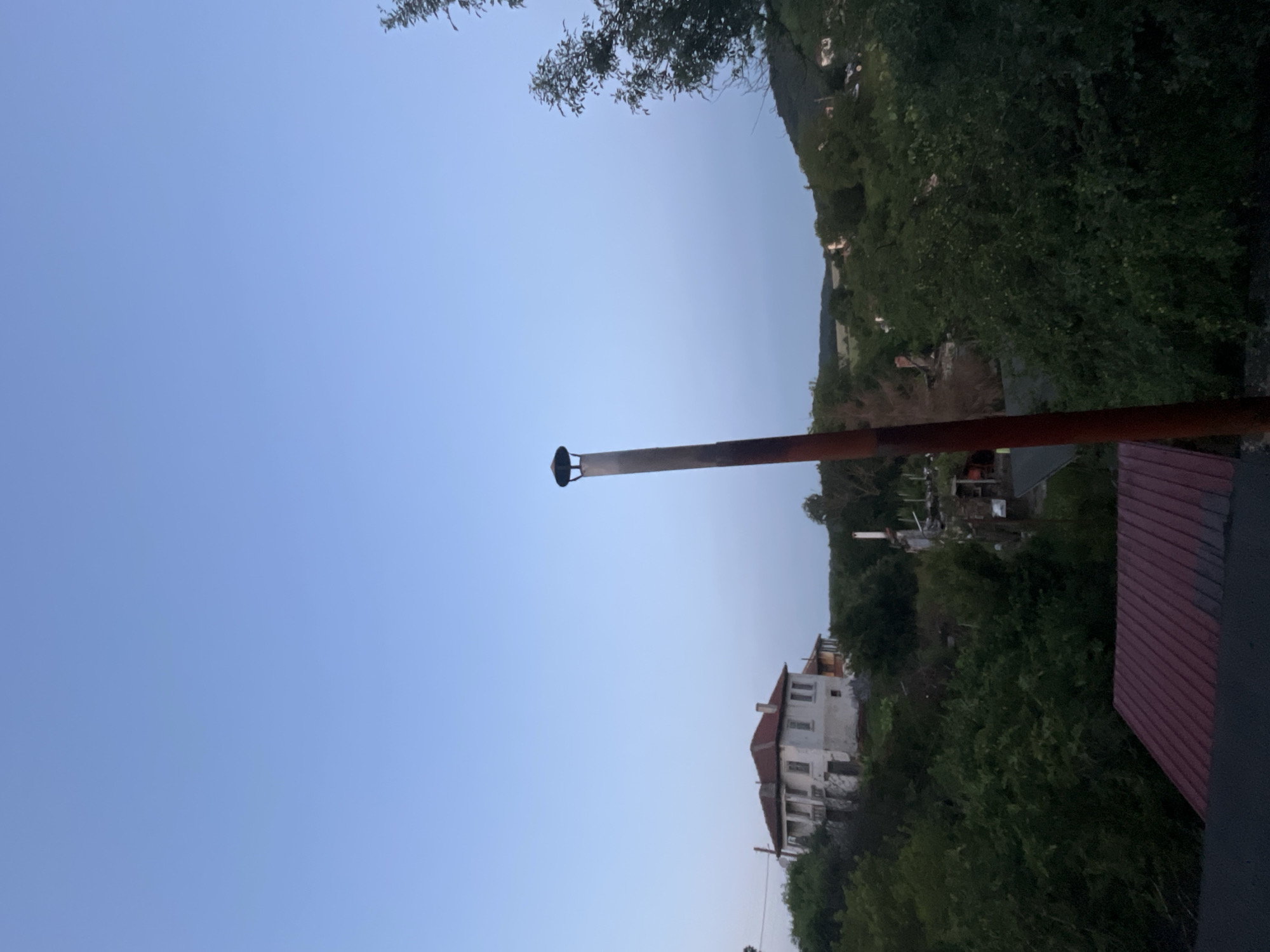 The image depicts a landscape identified as Varshilo, Bulgaria, during either dusk or dawn, characterized by a clear sky transitioning from a light blue overhead to a darker, almost purplish-blue near the horizon. Dominating the central-right portion of the frame is a tall, slender metal pipe, likely a chimney or vent, exhibiting a dark, weathered, possibly rusted surface and capped with a black, inverted-conical cover.
To the left, a prominent two-story, detached residential building with a reddish-brown tiled roof and light-colored, possibly white, walls is situated on a verdant hillside. This structure features multiple windows and a visible balcony. The surrounding landscape is densely covered with dark green foliage, comprising numerous trees and bushes that extend across the midground and into the background.
A portion of a dark red corrugated metal roof is visible in the lower-left quadrant of the foreground, suggesting another structure or property immediately below the vantage point. Further into the background, undulating terrain covered with more trees is discernible under the fading light. No individuals, animals, or specific actions are observed within the scene.