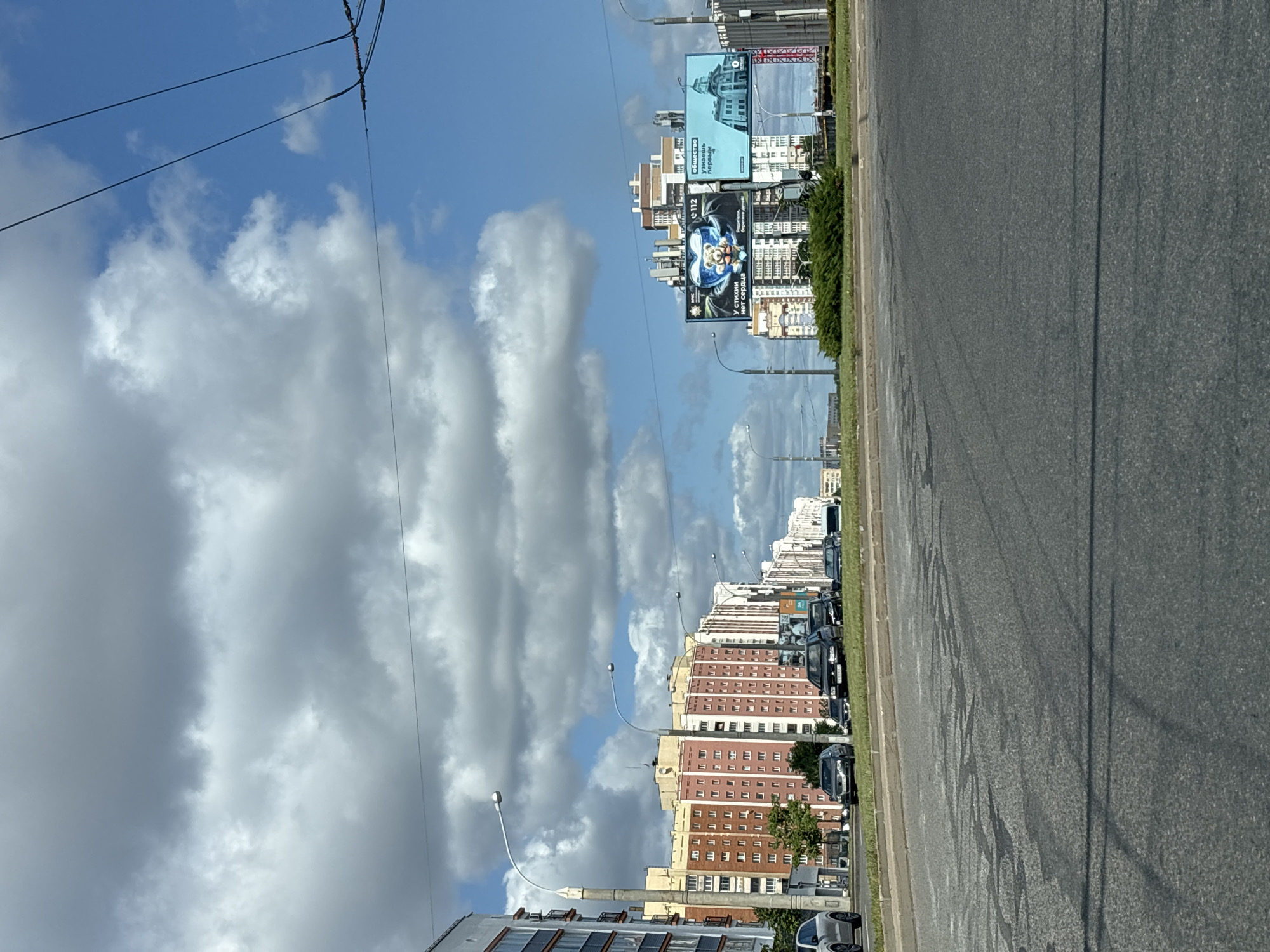 The image captures a street scene in Minsk, Belarus. In the foreground, an asphalt road is visible, with tire marks and dark patches indicating wetness. A few shadows stretch across the road, suggesting a sunny day with some cloud cover. The middle ground features a grassy median strip bordered by a curb. A line of cars is parked or driving on the road to the left.
Further back, a row of multi-story apartment buildings lines the street. These buildings have a mix of light and dark colored facades, with numerous windows. Streetlights with curved poles are positioned along the road.
On the right side of the image, a large billboard is mounted on a structure. The billboard displays advertisements, one of which features a teddy bear inside a heart shape and text in Cyrillic. Another part of the billboard shows a building illustration. More apartment buildings and some construction structures are visible in the distance.
The sky above is a vibrant blue, dotted with large, fluffy white and gray clouds, indicating a partly cloudy day. Overhead, power lines are strung across the sky, attached to poles. The overall impression is of a typical urban environment in a city like Minsk.