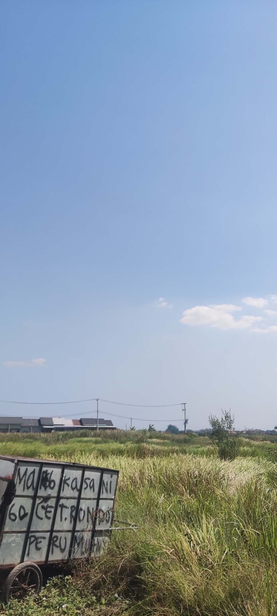 In Gebang, Indonesia, a weathered metal cart stands amidst tall, dry grass. The side of the cart is inscribed with black spray-painted text, some of which is legible: "MA KASA NI", "60 CET RONDO", and "PERU MUHLN". In the background, a low-rise building with a segmented roof and power lines stretching across the horizon are visible under a clear, pale blue sky. The environment suggests a rural or semi-rural setting with a significant amount of vegetation.