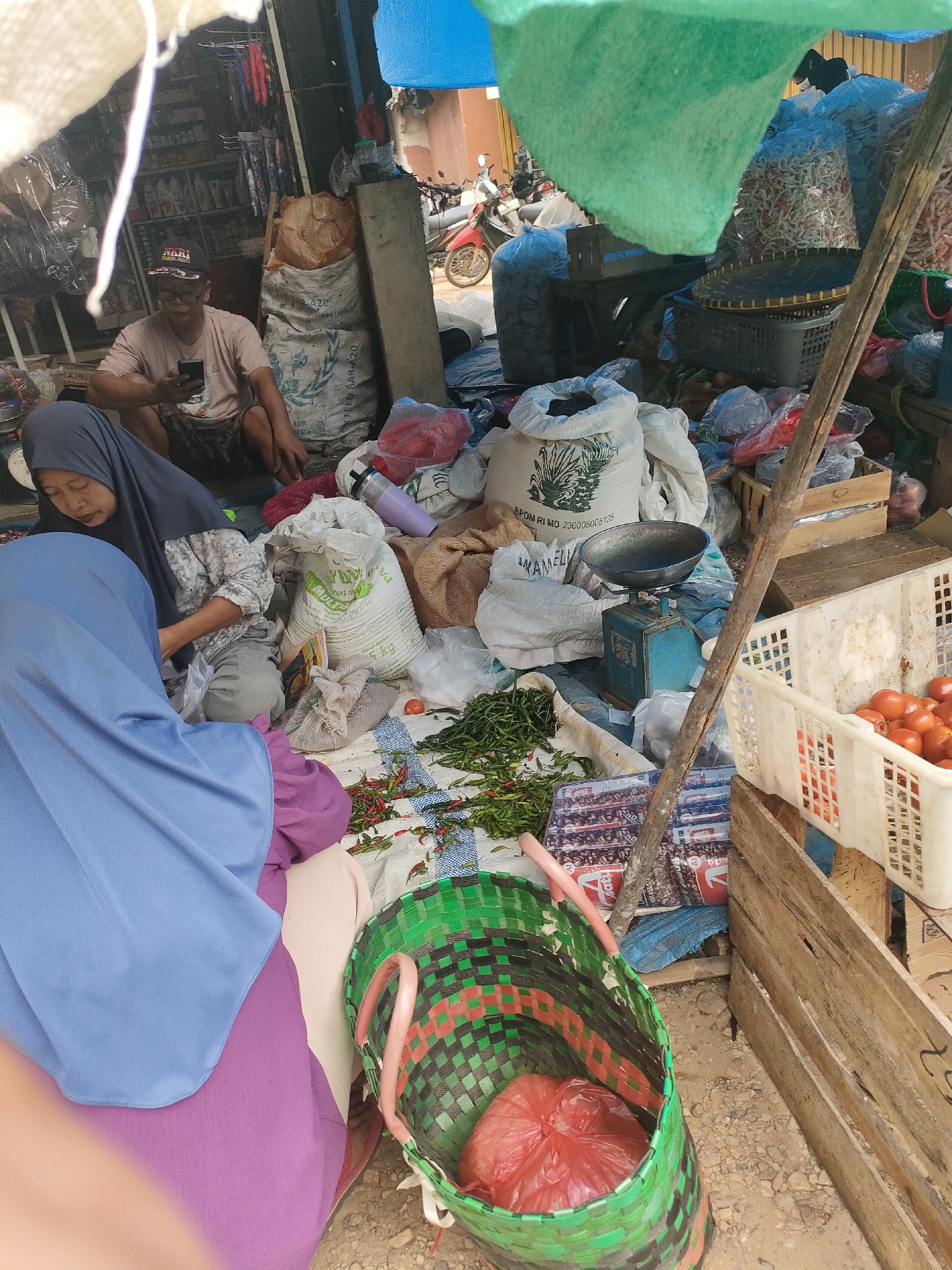 The image depicts an outdoor market scene in Sukoyoso, Indonesia. A man wearing a cap and a t-shirt is seated on the ground, looking at his mobile phone. Two women, both wearing hijabs, are also seated on the ground. One woman, in a dark grey hijab, is looking down and appears to be engaged in an activity. The other woman, in a light blue hijab, has her back to the camera, and her purple clothing is visible.
Various goods are spread around them, including sacks of what appears to be rice or grain, baskets filled with tomatoes and other produce, and a pile of red chili peppers. A weighing scale with a blue base and a metal pan is placed on the ground near some sacks. A green woven basket containing a red plastic bag is in the foreground. The market stall is covered by a blue and green tarp. In the background, a motorcycle is visible, suggesting the general environment of a bustling marketplace. The ground is uneven and appears to be dirt or gravel.