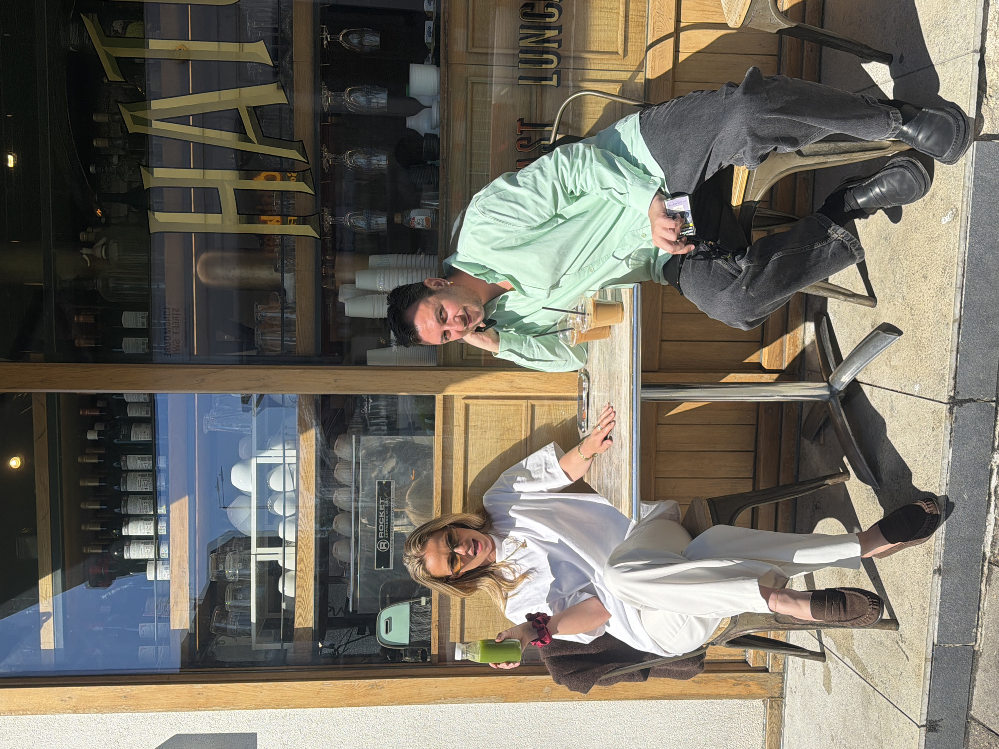 Two people sit at an outdoor café table on a sunny day. A man with dark hair wears a light green shirt and dark jeans. He sits in a chair and holds a small item in his right hand. Beside him on the table are two iced coffee drinks. Across from him, a woman with blonde hair wears a white outfit and holds a green juice bottle. They are outside a wooden-framed building, a café or restaurant, with a large window displaying wine bottles and coffee equipment. "BAR" is displayed on the windows, with "LUNCH" to the right. The scene appears to be in London, United Kingdom.