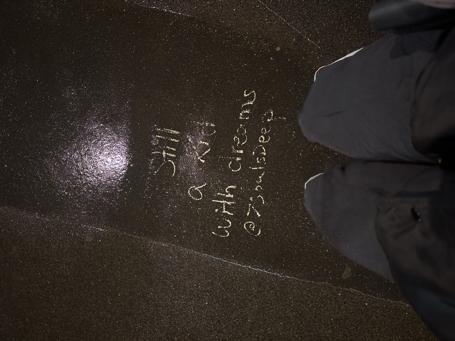 Chalk writing on dark pavement reads "Still a kid with dreams @7soulsDeep". Shiny pavement reflects light near the text. A person wearing dark pants and light-soled shoes stands to the right. A seam runs across the lower part of the pants. The overall tone is dark and reflective.
