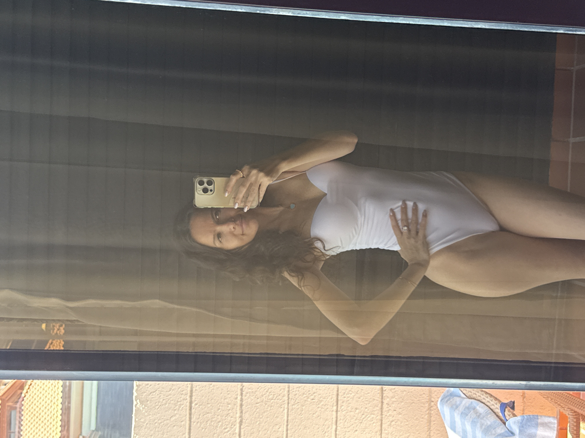 A woman is taking a mirror selfie. She is wearing a white strapless swimsuit. She has dark curly hair and is wearing a ring on her left hand. She is holding a smartphone to take the picture. The background appears to be an indoor setting with a large mirror, possibly in a hotel room. There is a glimpse of an outdoor area through a sheer curtain to the left of the mirror, showing a tiled roof and a chair with a striped towel. The lighting is natural and bright, suggesting it's daytime. There is no information in the image that can place the location in Dubai, United Arab Emirates.