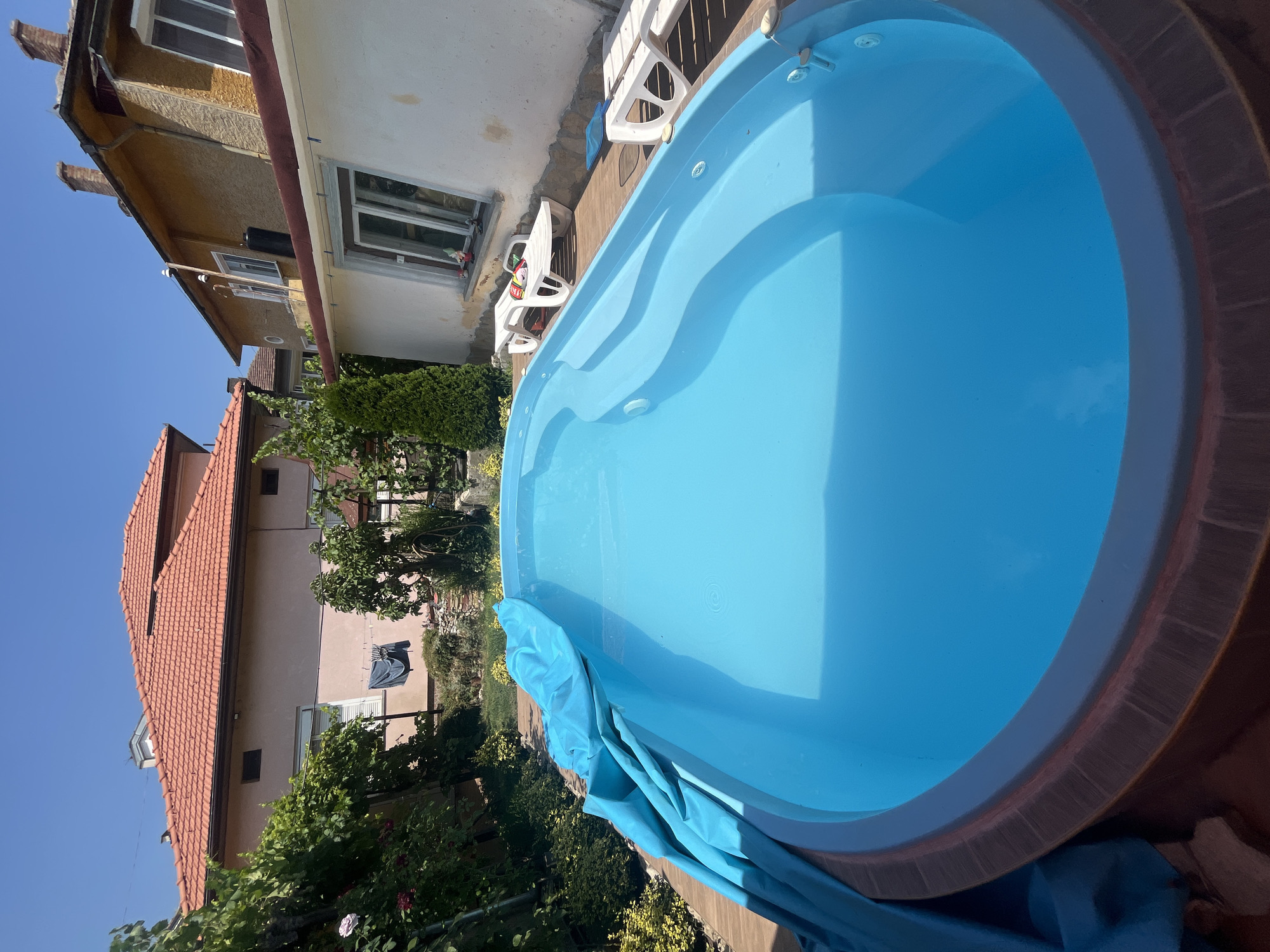 The image depicts an outdoor scene, likely a residential backyard, in Pavlikeni, Bulgaria, on a sunny day. In the foreground, a large, bright blue swimming pool is partially covered by a crumpled blue tarp. The pool appears to be empty or very shallow, with a wooden deck surrounding it.
In the middle ground, two white plastic lounge chairs are placed on the deck. One chair has a few items on it, including a striped ball and a colorful object. A blue pool skimmer lies on the deck near the second chair. Beyond the pool, two residential buildings are visible. The building on the left has a light pink facade and a red-tiled roof. Laundry, including a dark blue garment, is hanging on a line in front of this building. The building on the right is a yellow-colored structure with a red-tiled roof and a white section. Several windows are visible on both buildings.
The background consists of a clear blue sky and lush green vegetation, including trees and bushes, suggesting a warm climate. The overall impression is a summer scene in a private outdoor space.