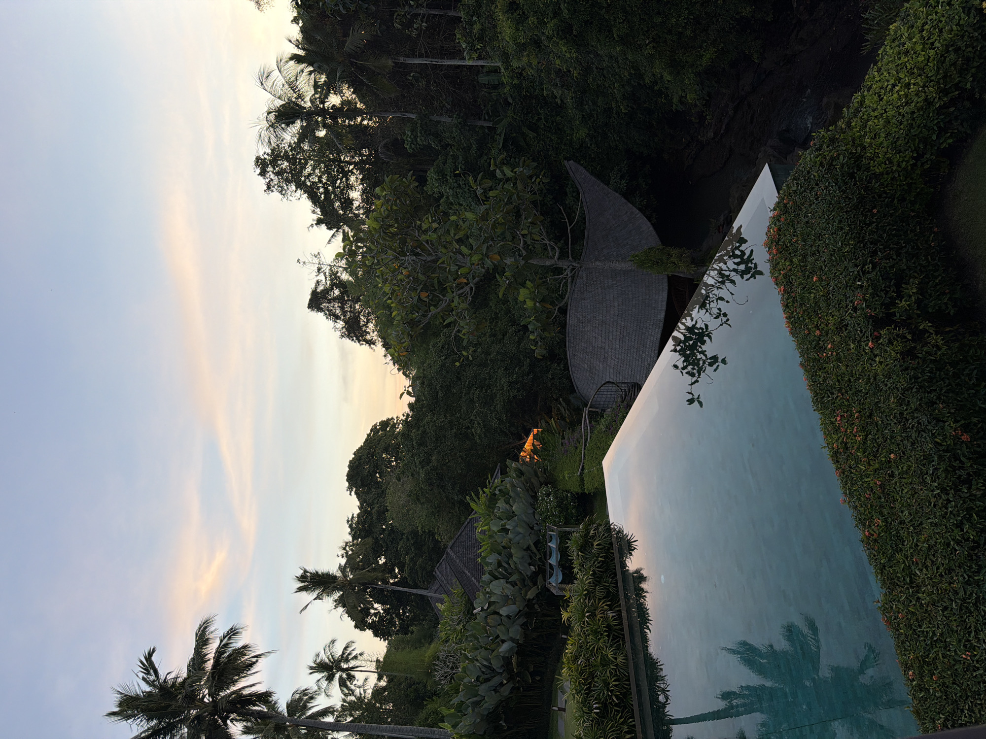 The image shows an outdoor scene at dusk, likely in a tropical or subtropical location. A swimming pool occupies a significant portion of the lower right, with its water reflecting the sky. Lush, dense greenery surrounds the pool, including various plants, bushes, and trees. Some trees appear to be palm trees. A building with a unique, curved roof is partially visible amidst the foliage. The sky is a mix of light blue and orange hues, suggesting sunset or sunrise.
No people are visible. The location is presumed to be near Kediri, Indonesia, although specific identifying features of the city are not discernable from the image alone.
