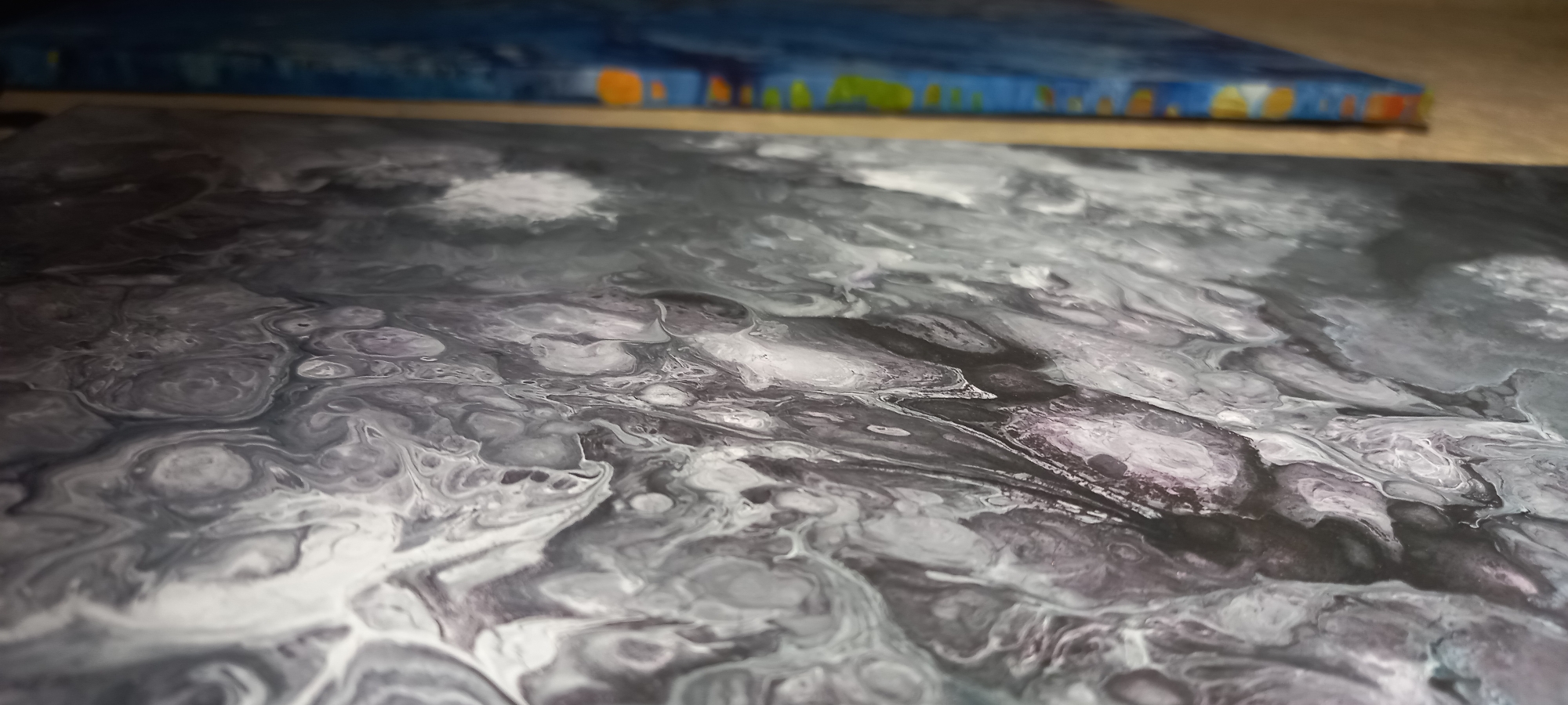 The image displays an abstract painting, viewed at a sharp upward angle, featuring fluid, swirling patterns of black, white, and various shades of gray, with faint undertones of purplish-grey. The paint exhibits characteristic lacing and cell formations typical of acrylic pour art. Dynamic curves and organic shapes dominate the composition, creating a sense of movement across the surface. Along the right edge, a narrow, light-colored wooden frame or canvas edge is visible, slightly out of focus. Behind this edge, a blurred blue surface with indistinct yellow and orange patches can be discerned. The environment appears to be an indoor setting. The image contains no identifiable landmarks, structures, or specific environmental cues that would allow for the determination of its location in Kryvyi Rih, Ukraine, or any other geographic area. No individuals or specific actions are depicted.