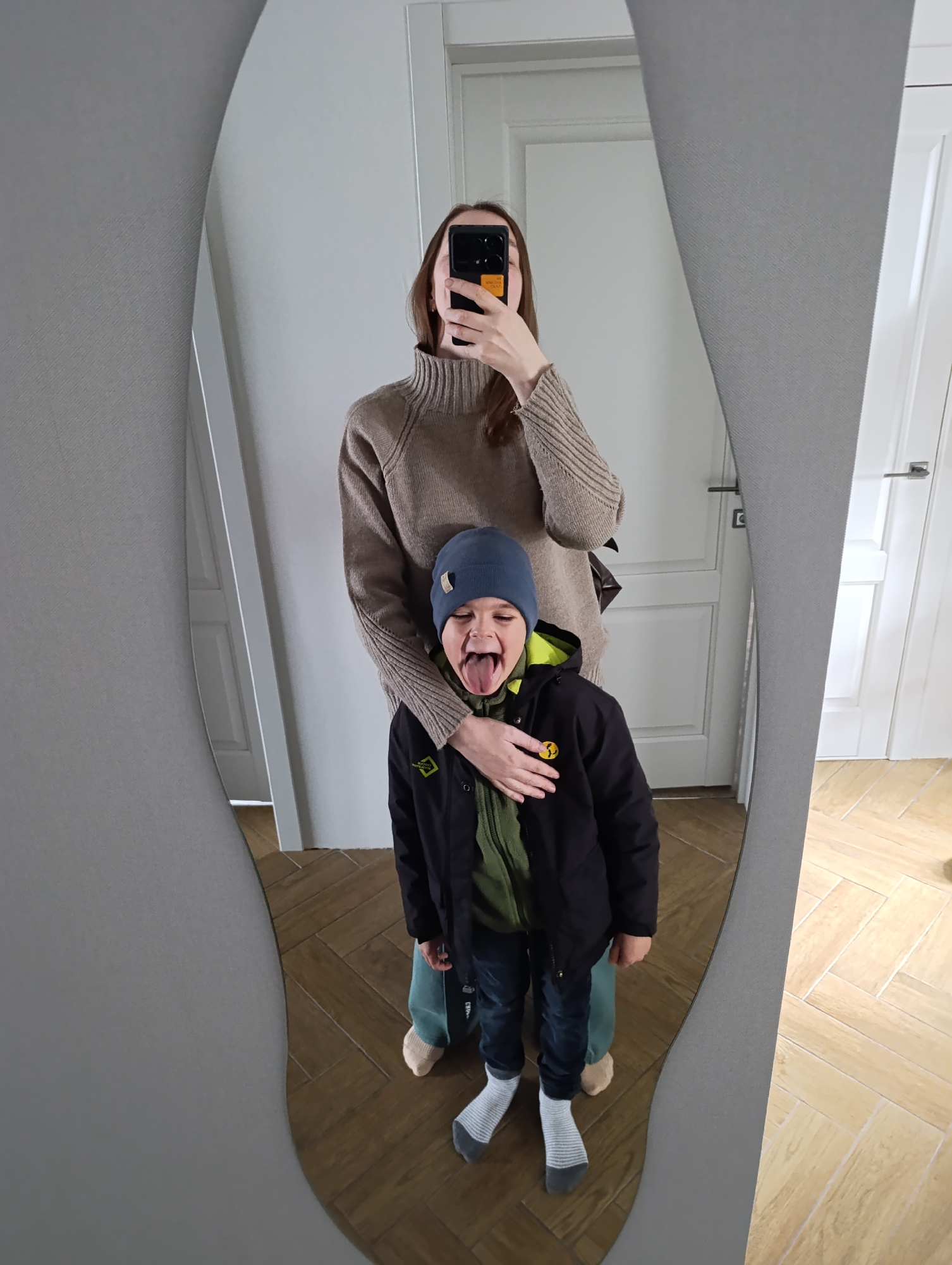A mother and child pose for a photograph in front of a mirror. The location is a room with light-colored walls and a tiled floor, in Chaikovsky, Russia. The mother, holding a phone, is wearing a beige turtleneck sweater. The child, wearing a blue beanie, a black jacket over a green garment, jeans, and mismatched socks, is sticking out their tongue. The mirror reflects a doorway and a partially visible second door.
