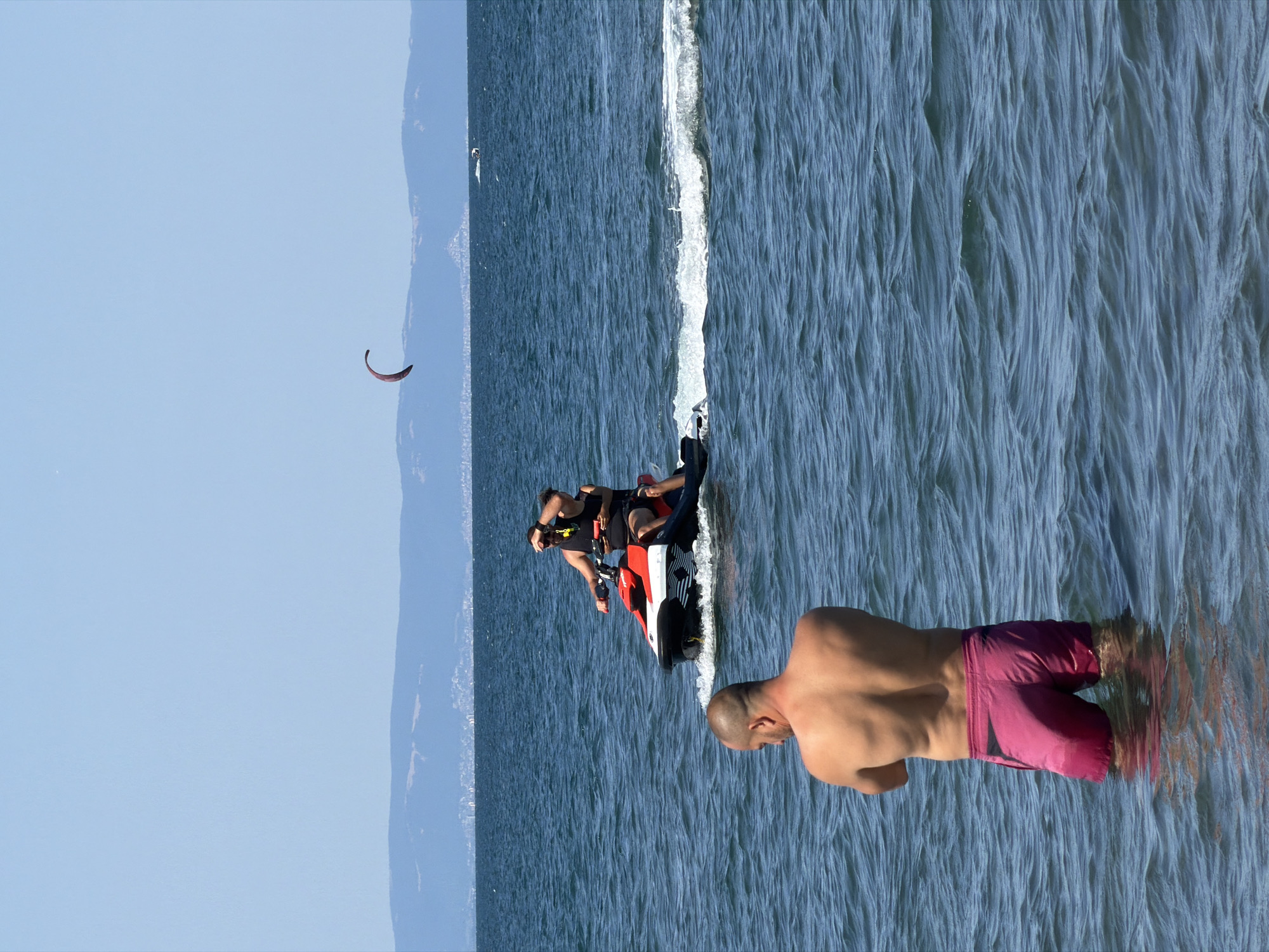 In the city of Atiya, Bulgaria, on a clear, sunny day, a man stands in the shallow, wavy, blue sea, facing away from the camera with his arms crossed. He is wearing pink swim trunks. Behind him, a jet ski is being ridden by two individuals, creating a wake. In the distance, a kite is visible in the sky over the ocean, and a hazy coastline with hills and buildings can be seen on the horizon.