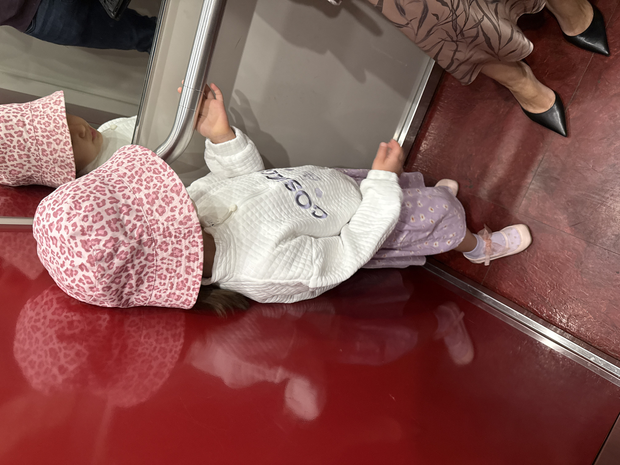 An overhead view captures a young child lying on a highly glossy, reflective red surface, likely a floor or bench, in an indoor public setting in Krasnoyarsk, Russia. The child, appearing to be a toddler, wears a pink leopard print bucket hat, a white quilted long-sleeved top with the word "COSIG" visible in purple letters across the chest, and light purple patterned pants. They also have on light pink mary-jane style shoes with white socks. Both of the child's hands are holding onto a silver metallic handrail or bar positioned along the left edge of the frame.
The reflective red surface prominently displays the child's reflection. In the upper right portion of the image, the lower legs and feet of an adult are visible, clad in a patterned dress or skirt and pointed black high-heeled shoes. A mirrored surface behind the child reflects vague shapes, suggesting the presence of other individuals or objects in the background. The scene depicts a quiet moment, possibly play or rest, with the child engaged with their immediate surroundings. The artificial lighting suggests an indoor environment, possibly a train carriage, metro, or public building.