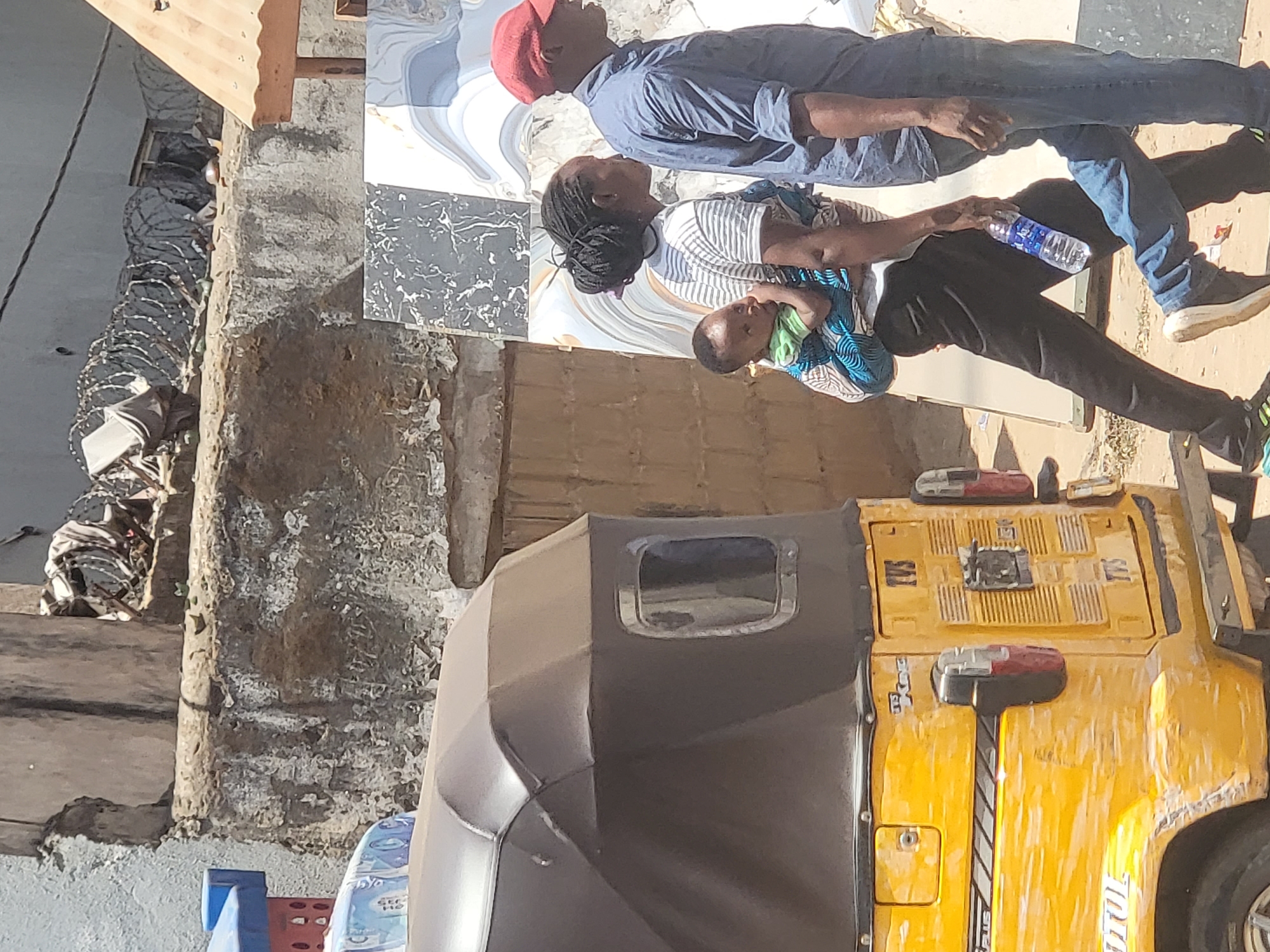 A yellow tuk-tuk, also known as a Keke Napep, is parked on a street in Maiburiji, Nigeria. Next to the tuk-tuk, three people are standing on the sidewalk; a man wearing a red hat, a woman carrying a baby in a sling, and another man next to them holding a water bottle. The walls of nearby buildings are visible. One is gray and appears to be made of concrete. A metal security barrier is attached to the top of this wall.