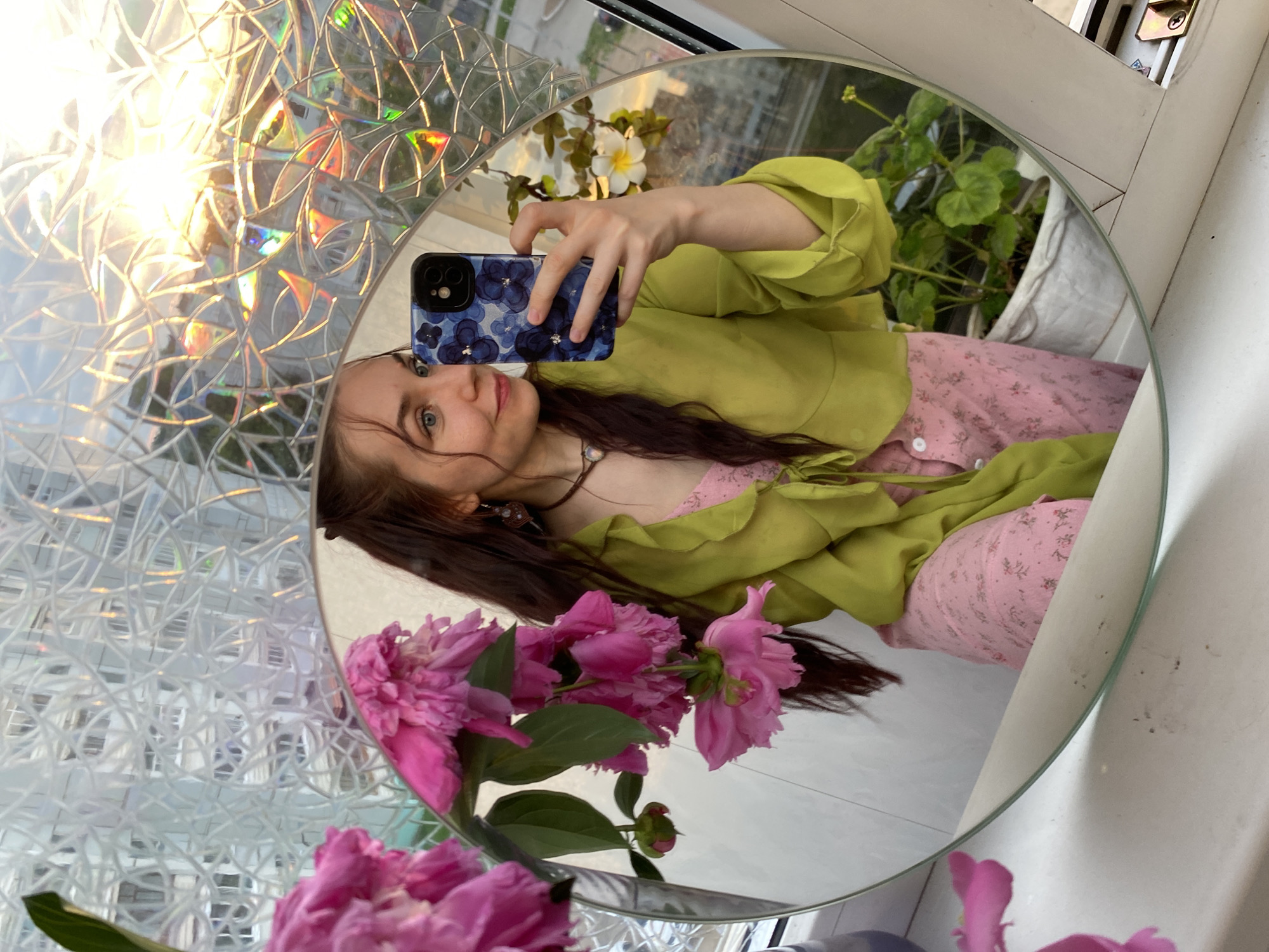 A young woman with long, dark hair is taking a selfie in a round mirror. She is wearing a lime green blouse tied at the waist and a pink floral print skirt or dress. In her right hand, she holds a smartphone with a blue floral case. Her left hand is visible near her face. She is looking to her right, with a slight smile.
The mirror reflects the woman, a bouquet of pink peonies in the foreground to the left, and a potted plant with green leaves and a white flower to the right. The background of the reflection shows a building with windows and a sunset, with light refracting through a patterned window covering. The scene appears to be indoors, likely near a window.
Based on the context provided, this image was taken in the city of Chaikovsky, Russia. The reflection of the building and sky suggests an urban setting.