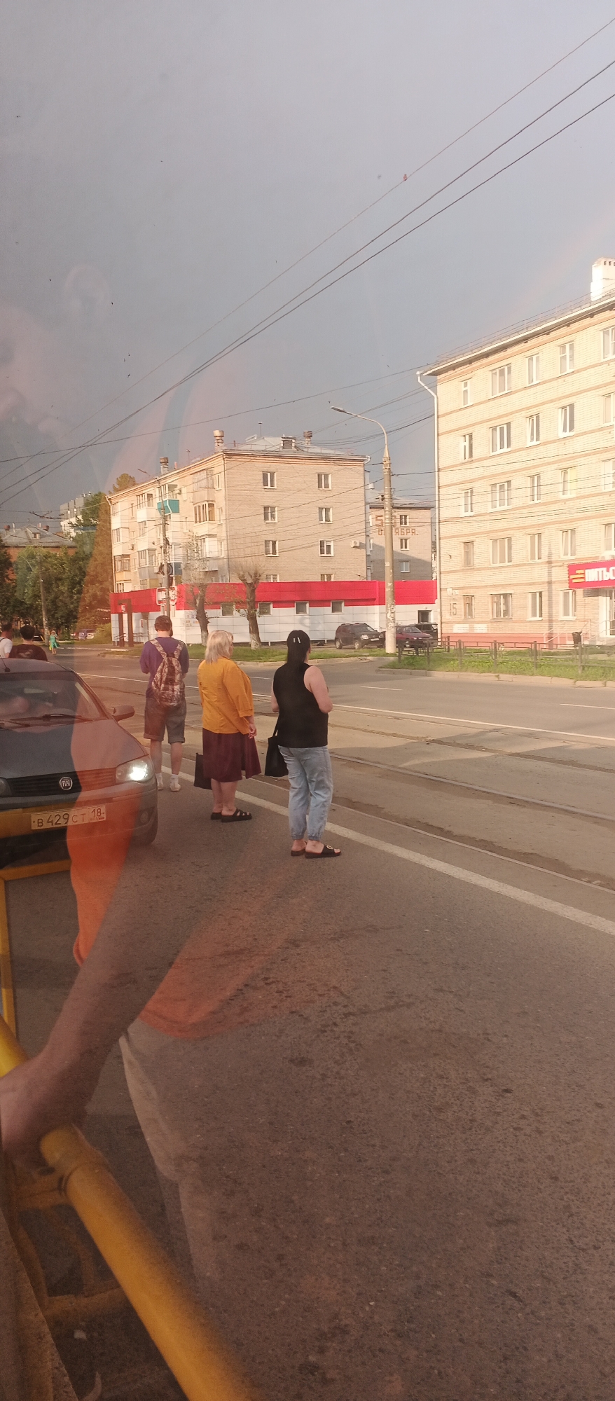 Three people are standing on a sidewalk next to a road in Izhevsk, Russia. The person on the left is wearing a purple shirt and shorts and has a backpack. The person in the middle is wearing a yellow shirt and a burgundy skirt. The person on the right is wearing a black sleeveless shirt and light blue pants. A car is parked on the left side of the road. In the background, there are several apartment buildings. The sky is overcast. The image was taken from inside a vehicle, as evidenced by the reflection on the left side of the frame.