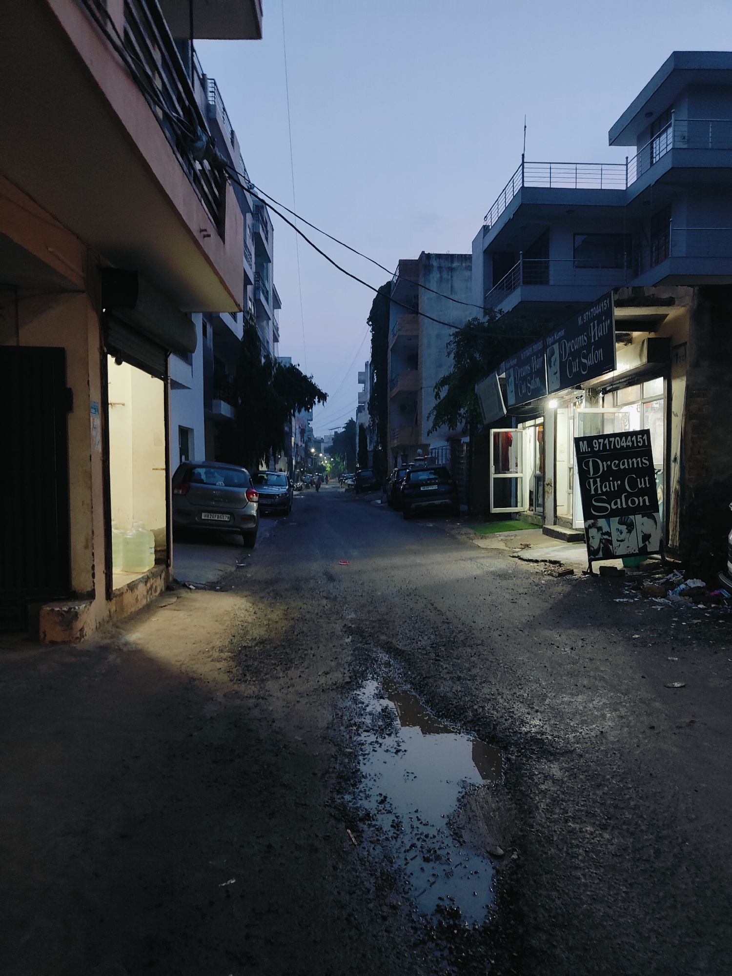 The image depicts a street scene at dusk in Gurgaon, India. The street is paved with asphalt and shows signs of disrepair, with potholes and standing water. On the right side of the street, a brightly lit storefront for "Dreams Hair Cut Salon" is visible, with a sign displaying a phone number. A portable sign in front of the salon shows three images of hairstyles. Several cars are parked along the street, and a few pedestrians can be seen in the distance. The buildings lining the street are multi-story structures, typical of urban Indian architecture. The sky is a muted blue, indicating twilight. The overall impression is of a typical residential or commercial street in a developing urban area during the evening hours.