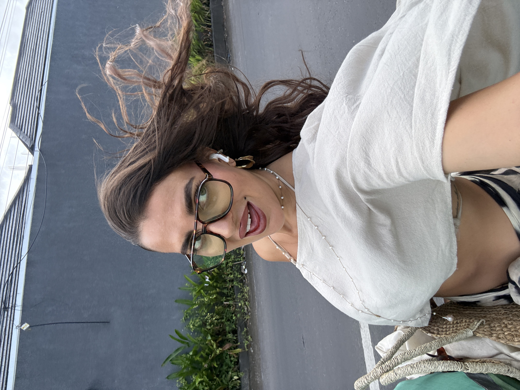 A woman with long brown hair, glasses, and a white top takes a selfie. Her hair is windblown. She wears jewelry, including earrings and a pearl necklace. A building and some green vegetation are visible in the background. A street or sidewalk is also visible. The location is Kuta Selatan, Indonesia.