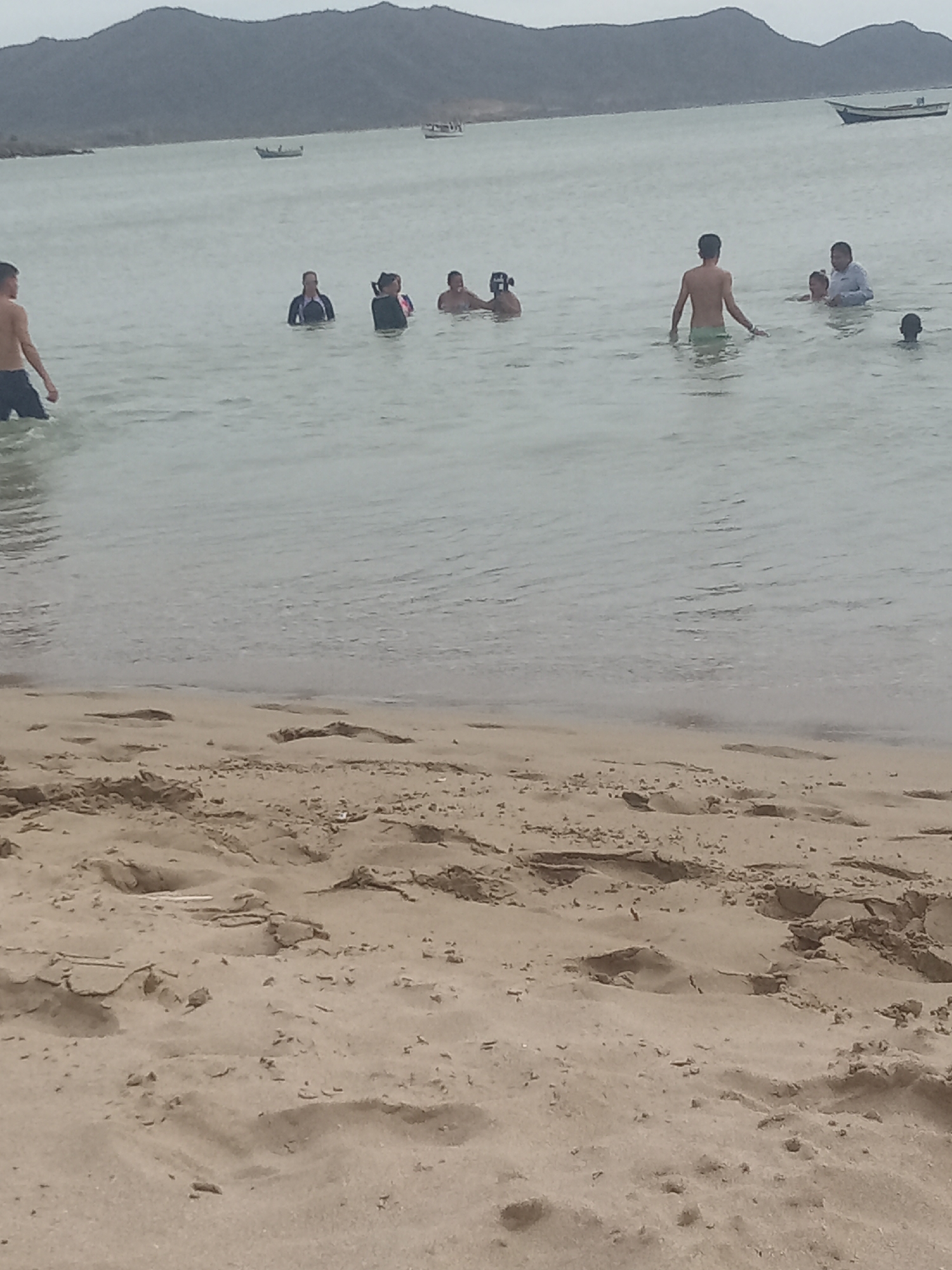 The image depicts a coastal scene in Juan Griego, Venezuela. A group of people are wading in the water at the edge of the beach. There are at least nine individuals of varying ages visible in the water. Some are standing or wading, while others are partially submerged. Further out on the water, there are small boats. In the background, there is a view of rolling hills. The foreground shows a sandy beach.