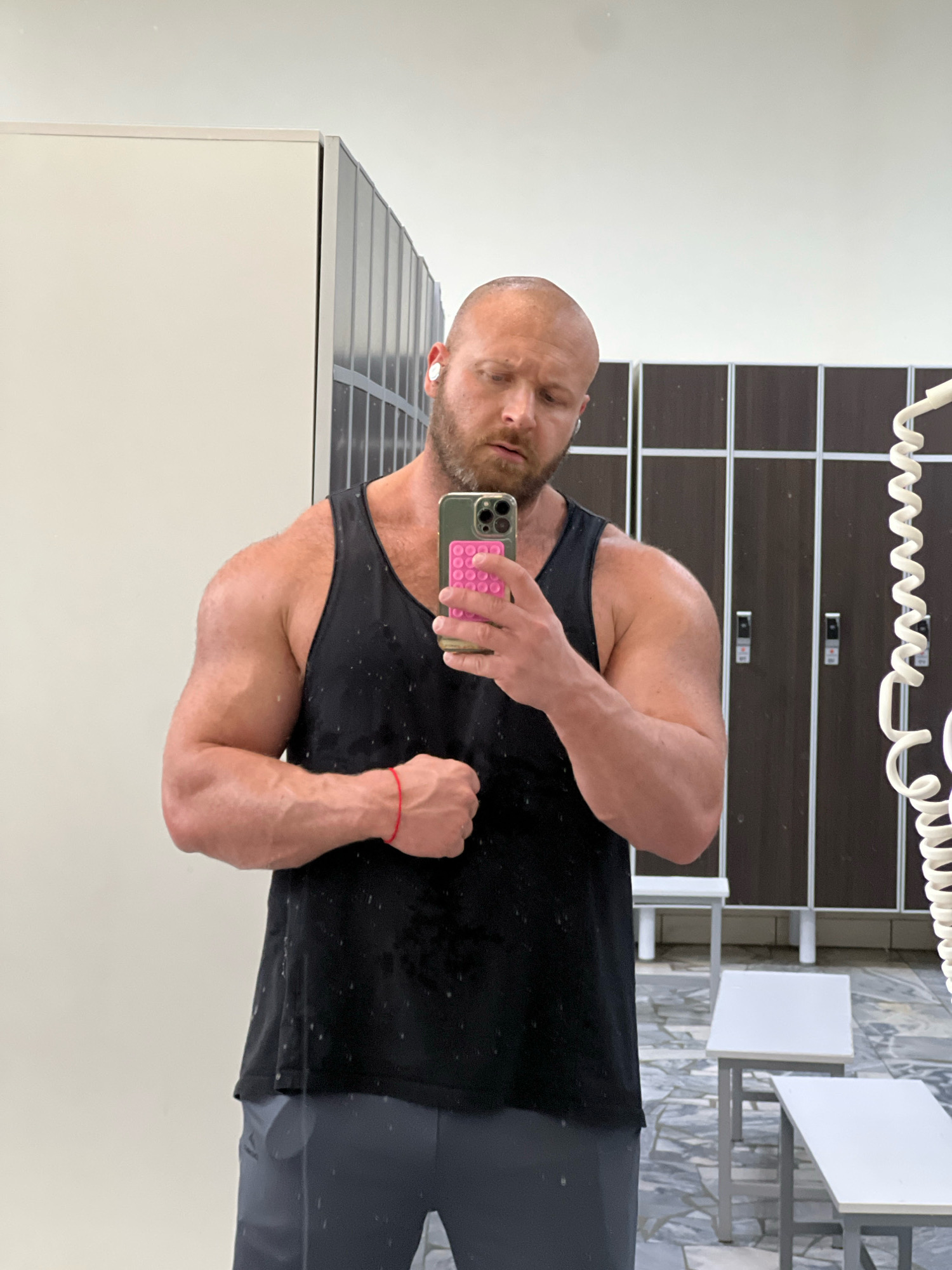A bald, white male with a beard is taking a selfie in a locker room. He is wearing a black tank top and grey athletic shorts. He has white earbuds in his ears and a red string bracelet on his left wrist. He is holding a green phone with a pink pop socket. The locker room has light-colored walls and dark-colored lockers. There are two white benches in the background. The floor is tiled with a grey and white pattern.