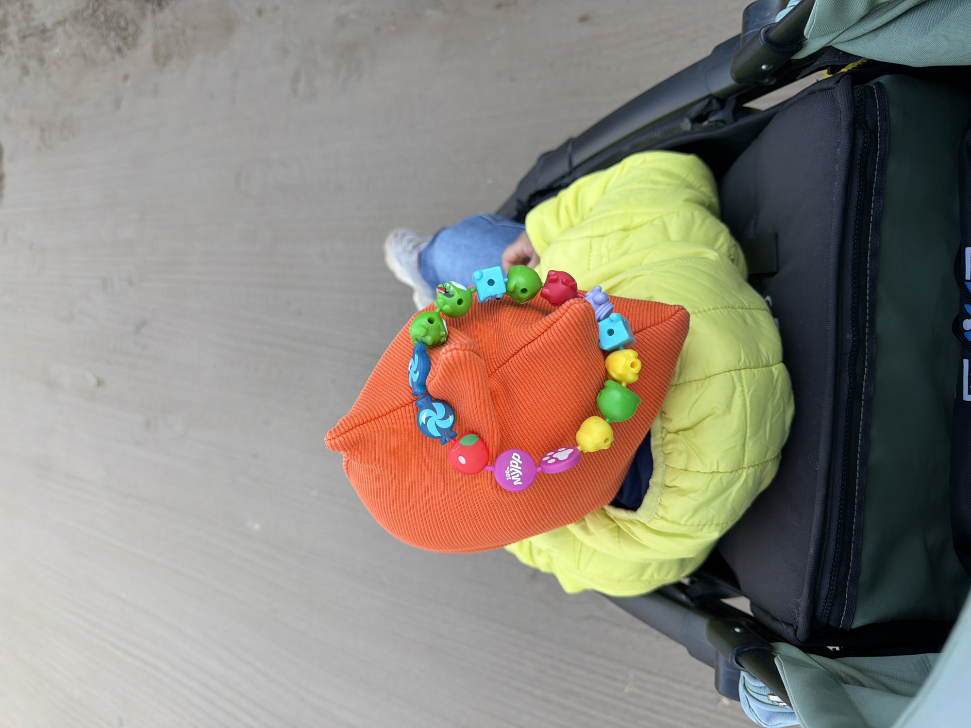A high-angle shot shows a stroller with a child inside, seemingly on a sidewalk. The child, dressed in jeans and a light-colored shoe, is partially covered by a bright yellow puffer jacket. An orange cap rests on top of the jacket. A colorful beaded toy adorns the cap. The stroller itself is black with hints of green and blue in the background. The sidewalk is made of gray concrete.
