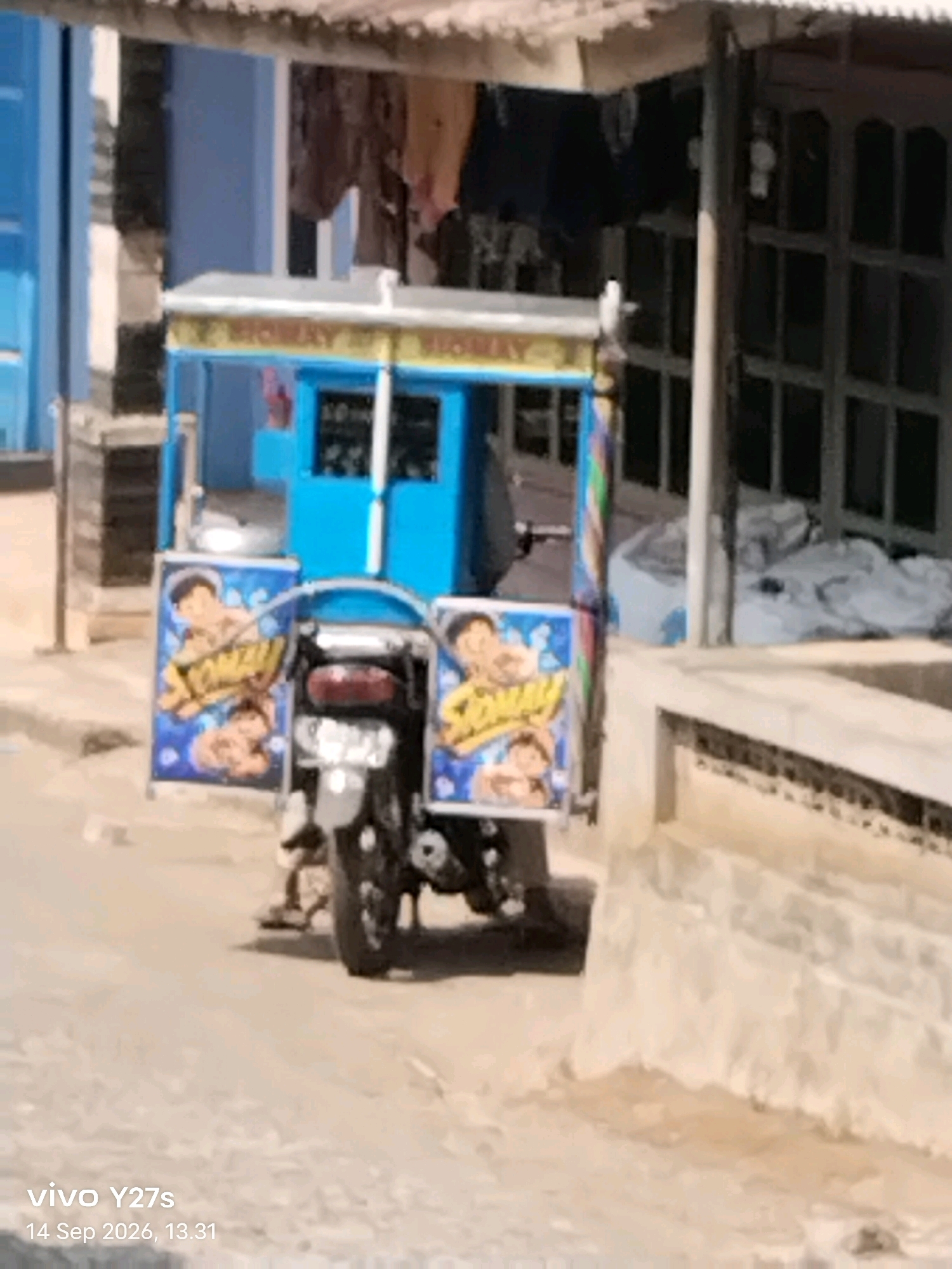 In Tamansari, Indonesia, a three-wheeled vehicle, commonly known as a bemo or trishaw, is parked on a paved street. The vehicle is painted bright blue and yellow, with decorative panels on its sides. These panels feature cartoon-like drawings of children with the word "SOMAY" written in yellow lettering. The front of the vehicle has a canopy, and it appears to be a mobile food cart, likely selling somay, a popular Indonesian street food. The background shows a building with blue doors and windows, and a corrugated metal roof. The overall scene depicts a typical street view in an Indonesian town.