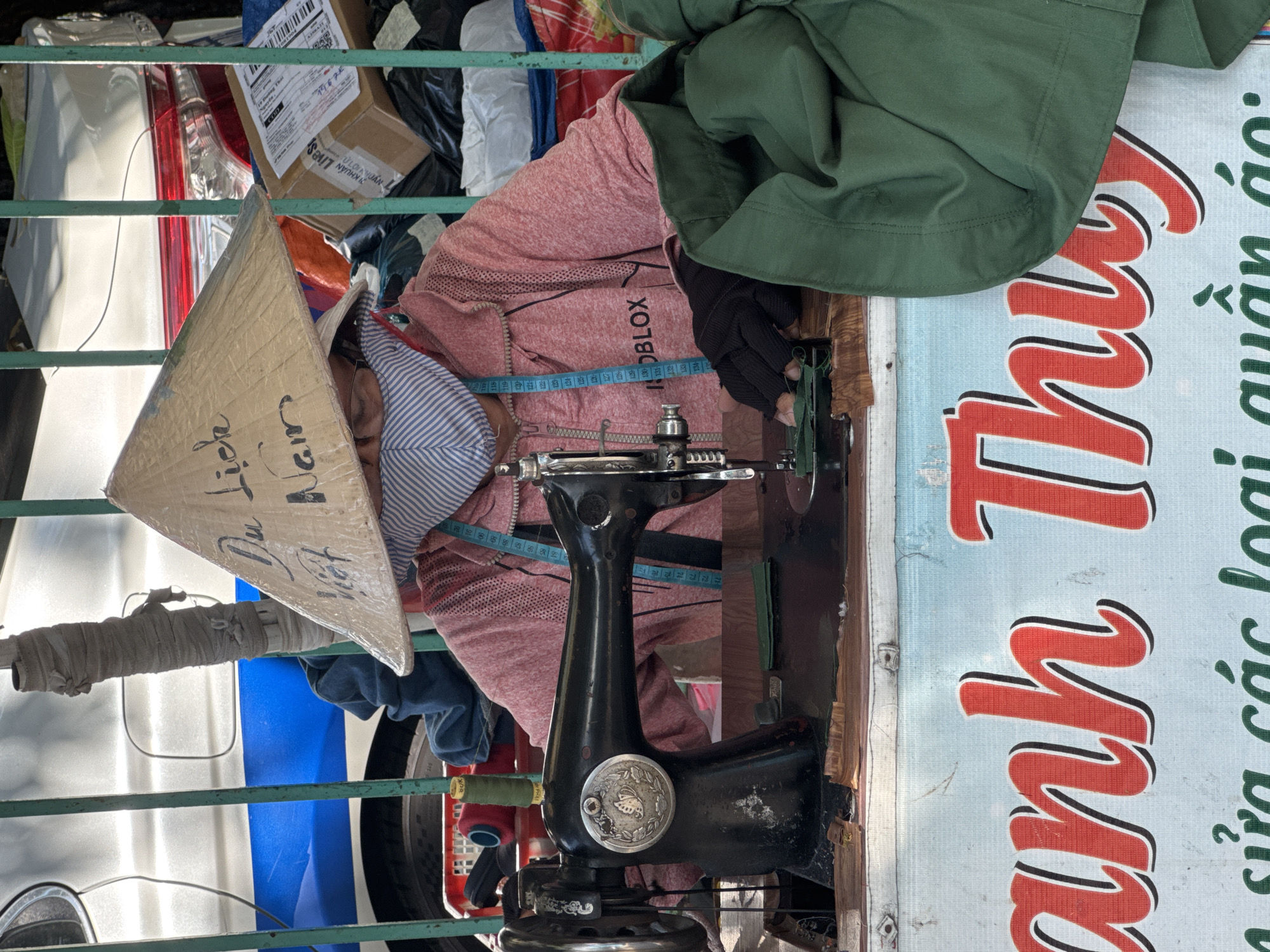 This media file captures a close-up view of a person engaged in sewing, likely in Nha Trang, Vietnam. The subject, partially obscured, is wearing a pink hooded jacket and black gloves, with a blue measuring tape draped around their neck. A traditional Vietnamese conical hat, known as a "nón lá," is placed on the head, with handwritten text on its surface reading "Du Lịch Wet Nam" (likely a misspelling of "Du Lịch Việt Nam," meaning "Vietnam Tourism").
The activity centers around a vintage black sewing machine, its intricate details visible. The background is a jumble of materials, including fabrics, boxes, and a glimpse of a white vehicle. A green cloth is draped over the shoulder of the person. To the right, a large sign with bold red lettering and black outlines is partially visible, displaying Vietnamese text that reads "anh Thu" and "sửa các loại quần áo:", translating to "Anh Thu" and "repairs all kinds of clothes:".
The scene suggests a workshop or a stall where sewing services are offered, possibly on a street or in a market. The presence of the conical hat and the Vietnamese text strongly indicates the location as Vietnam. The lighting suggests it could be daytime. The overall impression is one of industriousness and traditional craftsmanship.