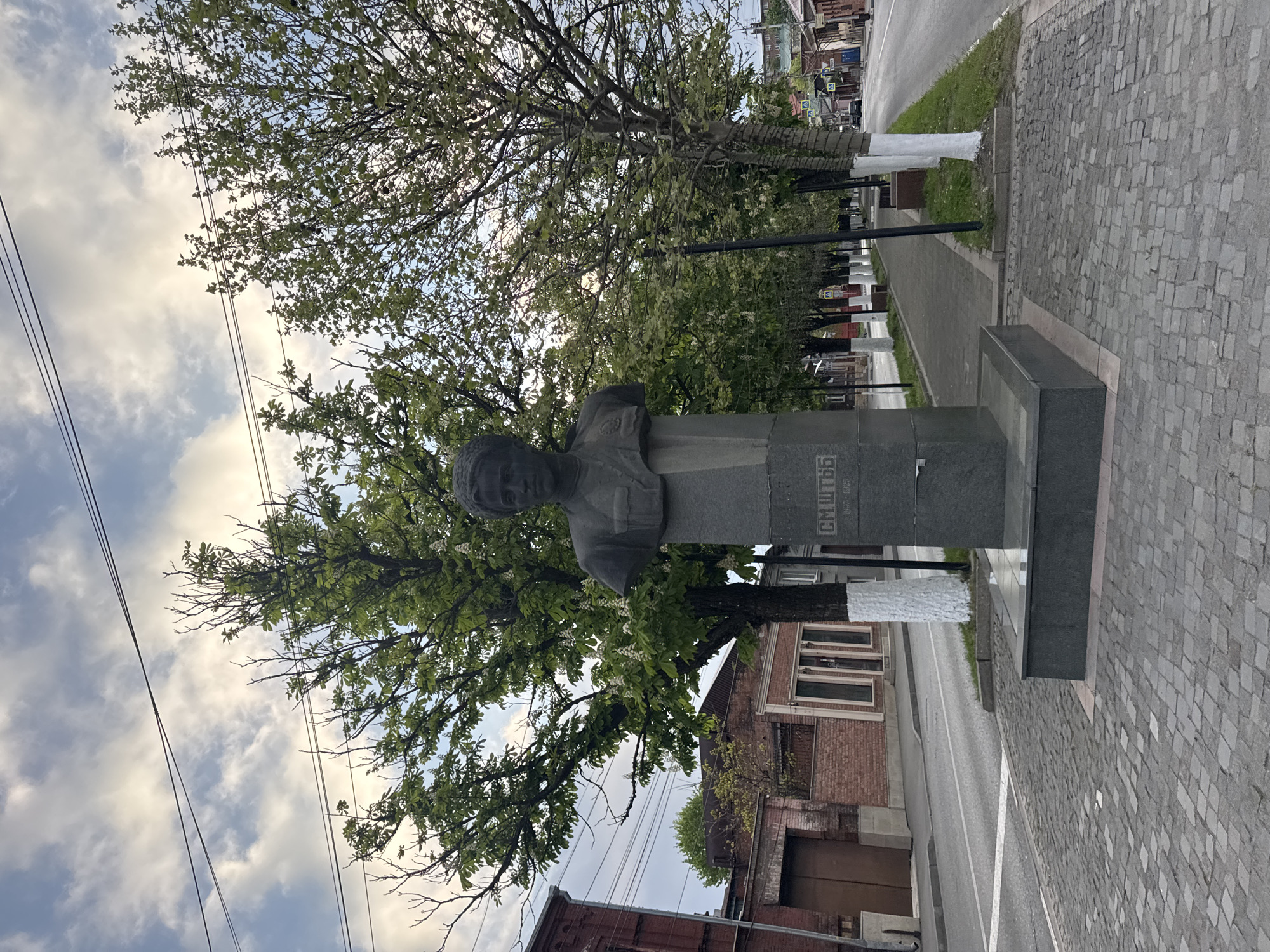 The image captures a view of a street scene in the city of Akhieli, Georgia. A stone bust of a man stands prominently on a plinth. The monument is situated along a sidewalk paved with interlocking stones. Behind the monument and sidewalk are trees, lining the pedestrian walkway, some of which are painted white at the base. A brick building is adjacent to the walkway. In the distance, other buildings can be seen along the road. The sky is visible above the buildings and trees.