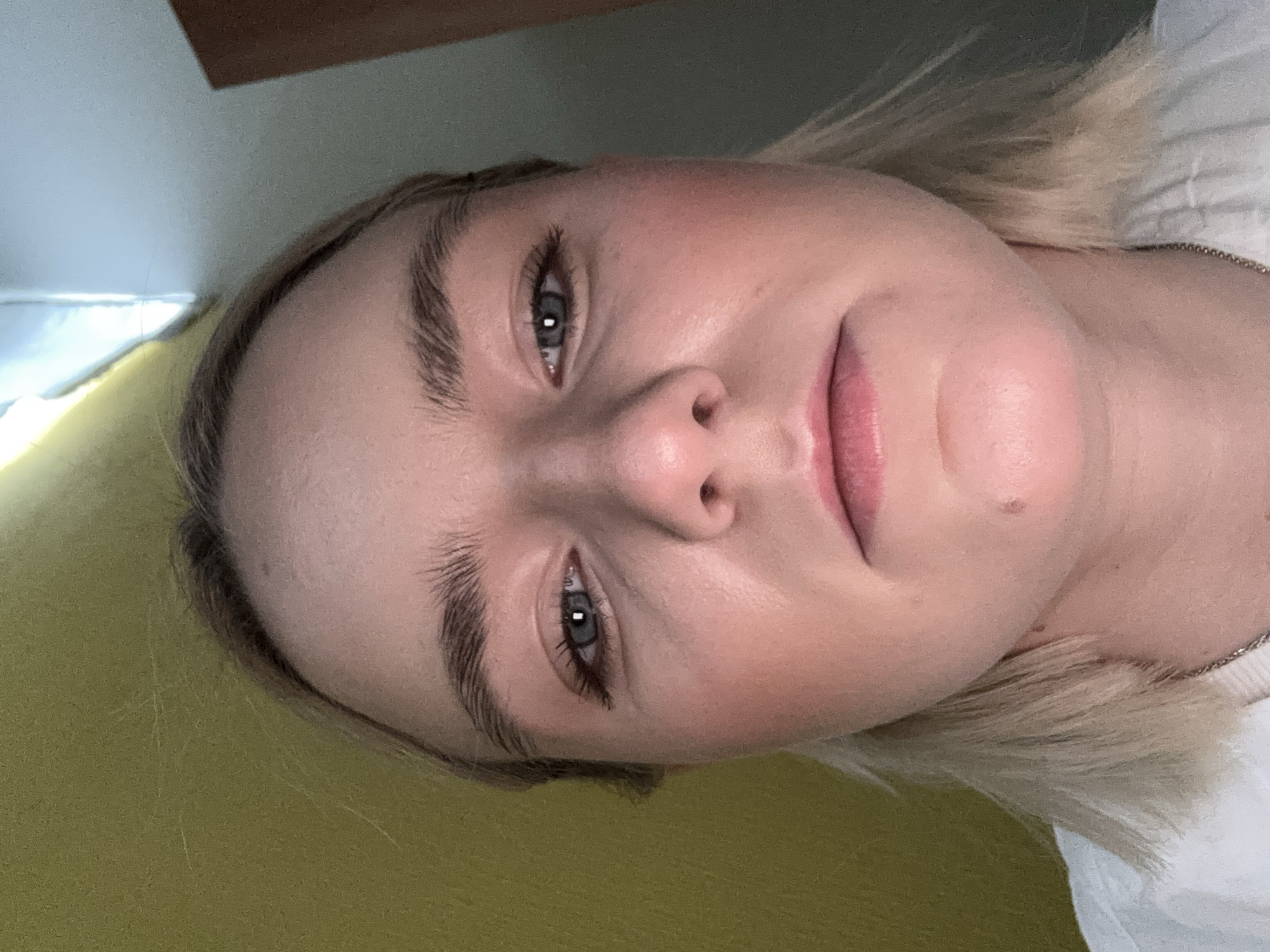 A close-up, slightly angled, portrait of a fair-skinned individual with blonde hair. The person has prominent eyebrows, blue/gray eyes, and pink lips. The background is blurred, featuring a light green/yellow surface on the left and a white object on the right. No location in Saint Petersburg, Russia, is discernible in the image.
