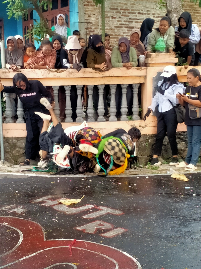 The image displays an outdoor scene in Jenggrik, Indonesia, featuring a cultural performance on a wet paved road.
**Environment:** The foreground is a dark, wet asphalt surface with partially visible red and white painted letters "HUT RI". Scattered debris, including yellow leaves and small paper fragments, is present. A grey concrete curb separates the road from a raised platform fronted by an orange-colored concrete balustrade with grey decorative pillars. Behind this platform, parts of a building with a blue facade and an exposed brick wall are visible, along with green foliage.
**People:**
Approximately 15-20 spectators, predominantly adult females and some children, are observing the scene from behind the balustrade, leaning forward and looking intently downwards. Their attire includes various headscarves (hijabs).
At ground level, on the left, an individual wearing a black long-sleeved shirt, black pants, and glasses is leaning against a grey pole. This person is actively engaged with three performers on the wet asphalt.
The performers are dressed in elaborate, colorful, and patterned traditional costumes, including masks and headpieces, and white athletic shoes. They are in a dynamic, prone, or tumbling position on the ground; one performer is partially upside down.
To the right at ground level, two adult females are standing. One wears a white baseball cap, a light blue long-sleeved shirt, black pants, and black gloves, holding a mobile phone and looking downwards. The other wears a black t-shirt, blue jeans, and light sandals, also holding a mobile phone and smiling.