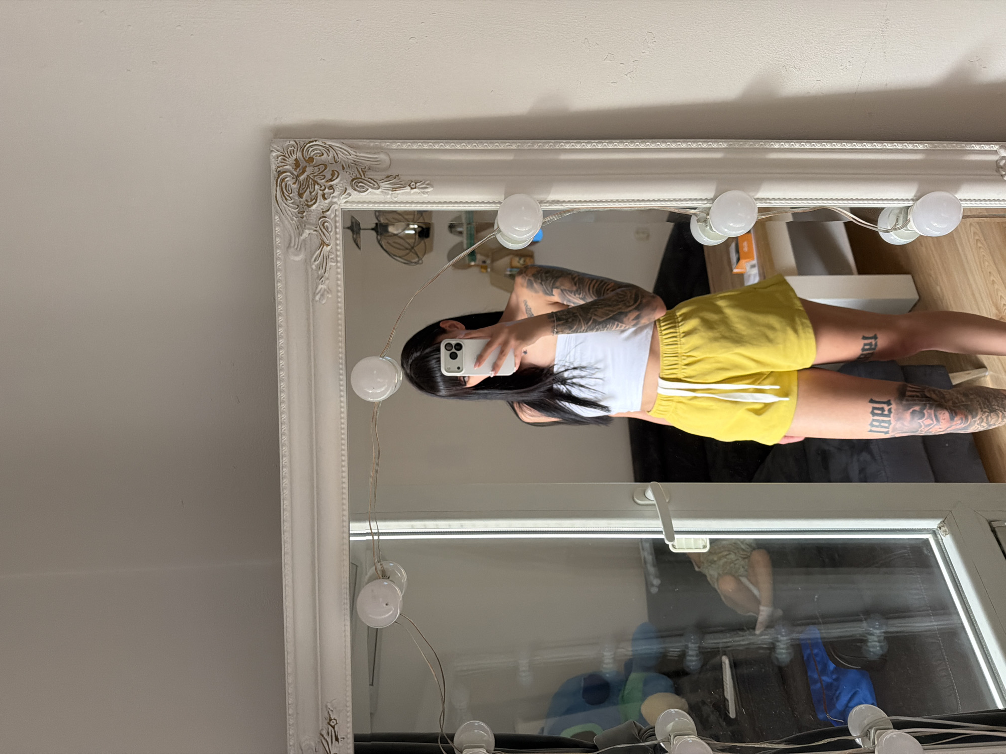 The image shows a young woman with extensive tattoos on her arms and legs taking a selfie in a well-lit mirror. She is wearing a white crop top and yellow shorts. The mirror is ornate with a white, carved frame and is surrounded by several small light bulbs, suggesting it might be a vanity or makeup mirror. The lighting is bright and even, indicating an indoor setting, likely a room in a house. In the reflection of the mirror, other objects can be seen, including furniture and what appears to be a window or glass door. There are also reflections of the light bulbs themselves. The overall impression is one of a casual, personal moment captured for social media. The visible text on the woman's leg is a large, stylized tattoo, but it is too blurry to read. The location context is Plovdiv, Bulgaria, but there are no specific visual cues in the image to confirm this. The time of day and weather are not discernible.