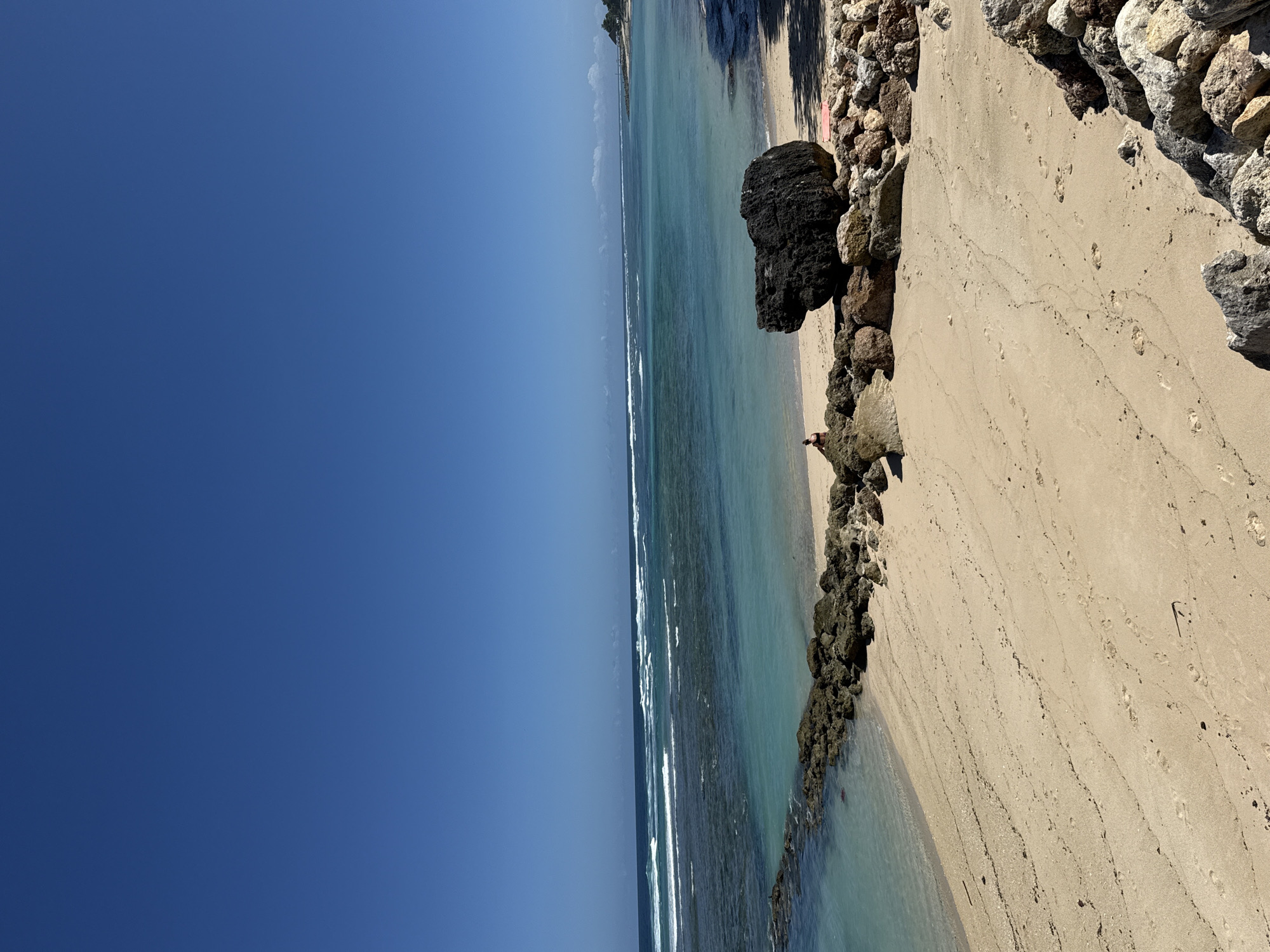 The image presents a view of a beach along the Kuta Selatan coastline of Indonesia. The scene is divided between a clear, bright blue sky and the ocean meeting the sandy beach. The water transitions from a light turquoise near the shore to a deeper blue further out. Waves are visible breaking along the reef line.
On the right side of the image, a sandy beach is bordered by a rocky barrier made of dark gray stones. Footprints are apparent in the sand. A large, dark-colored rock sits prominently on the beach.
A person is visible standing near the water's edge close to the rock structure. The general location is the coastline in Kuta Selatan, known for its beaches.