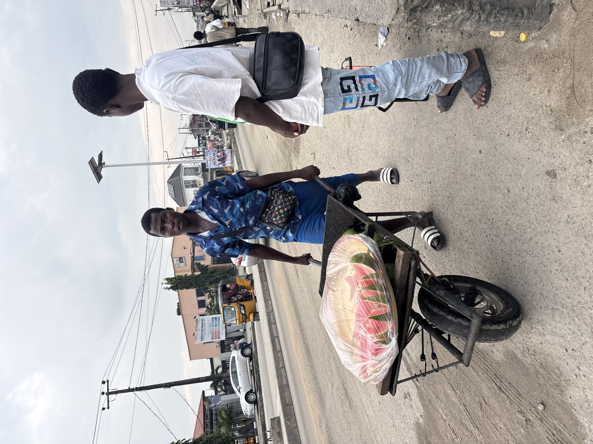 Two young men stand on a dusty roadside in Ado, Nigeria. The man on the left, facing the camera, wears a blue and black patterned short-sleeved shirt and blue shorts. He is pushing a wheelbarrow filled with sliced watermelons covered in plastic wrap. He wears white slides. The man on the right, facing away from the camera and towards the left, wears a white t-shirt and light blue jeans with a graphic design. He has a black crossbody bag and wears black slides. The background shows a street with a sidewalk, vehicles including a yellow auto-rickshaw, buildings, power lines, and a cloudy sky. The scene appears to be a marketplace or a busy street in a city.