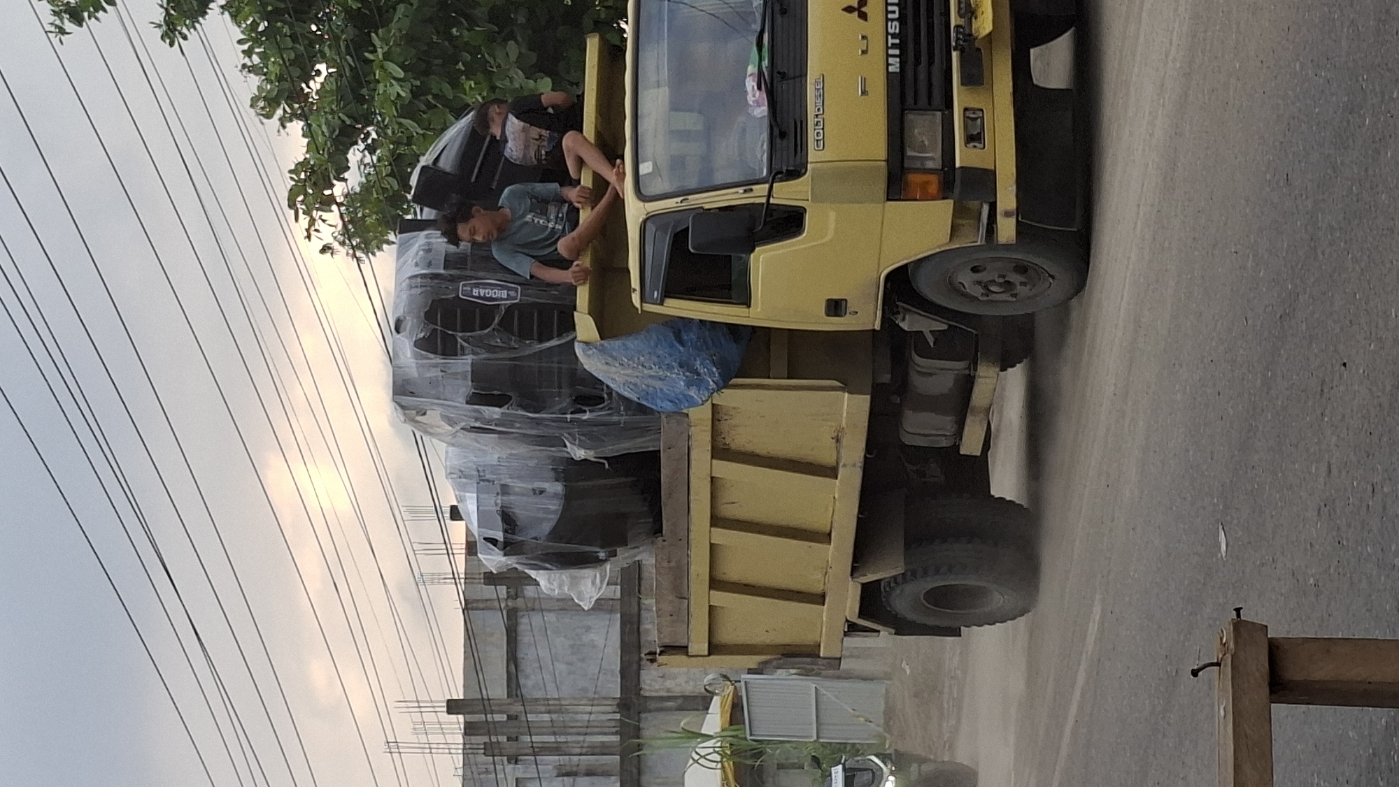 In Keudebayu, Indonesia, a yellow Mitsubishi Fuso dump truck is traveling on a paved road, kicking up dust. Two young males are seated on top of a large, black, speaker-like object covered in clear plastic wrap, which is loaded in the truck bed. Next to the speaker, a blue tarp is also visible. The truck's front features a "Colt Diesel" badge. A partially obscured building with rebar protrusions and trees are in the background. The sky is overcast with visible power lines overhead. A motorcycle is partially visible on the left side of the frame.
