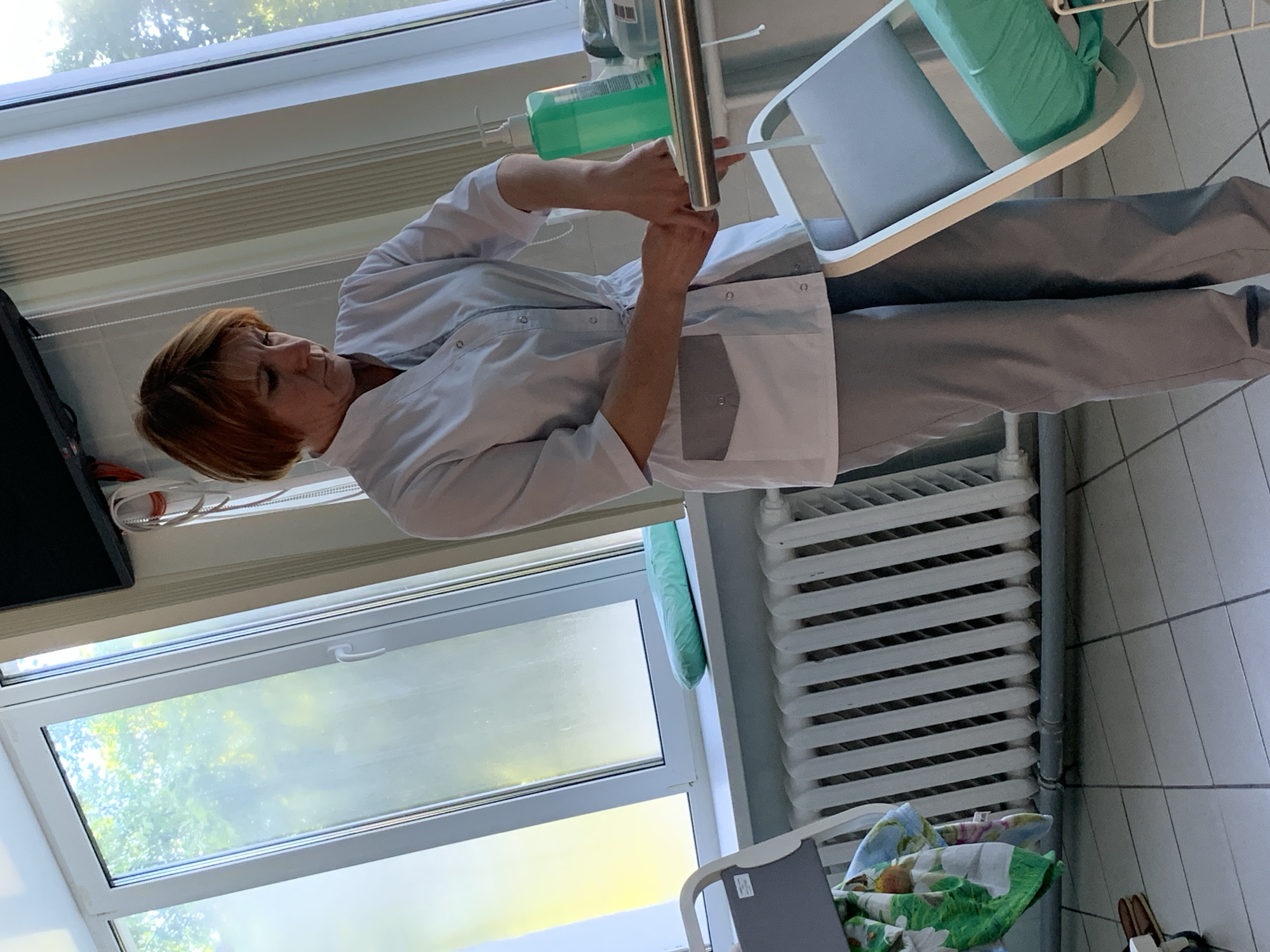 A Caucasian woman with short auburn hair, wearing a white medical coat with grey accents and grey pants, is seen from the waist down, indoors. Her hands are positioned as if washing them, with a green pump bottle of liquid nearby on a metal tray. The setting appears to be a medical facility, indicated by a radiator below a window, a medical cart, and tiled flooring. Outside the window, trees and sunlight are visible.