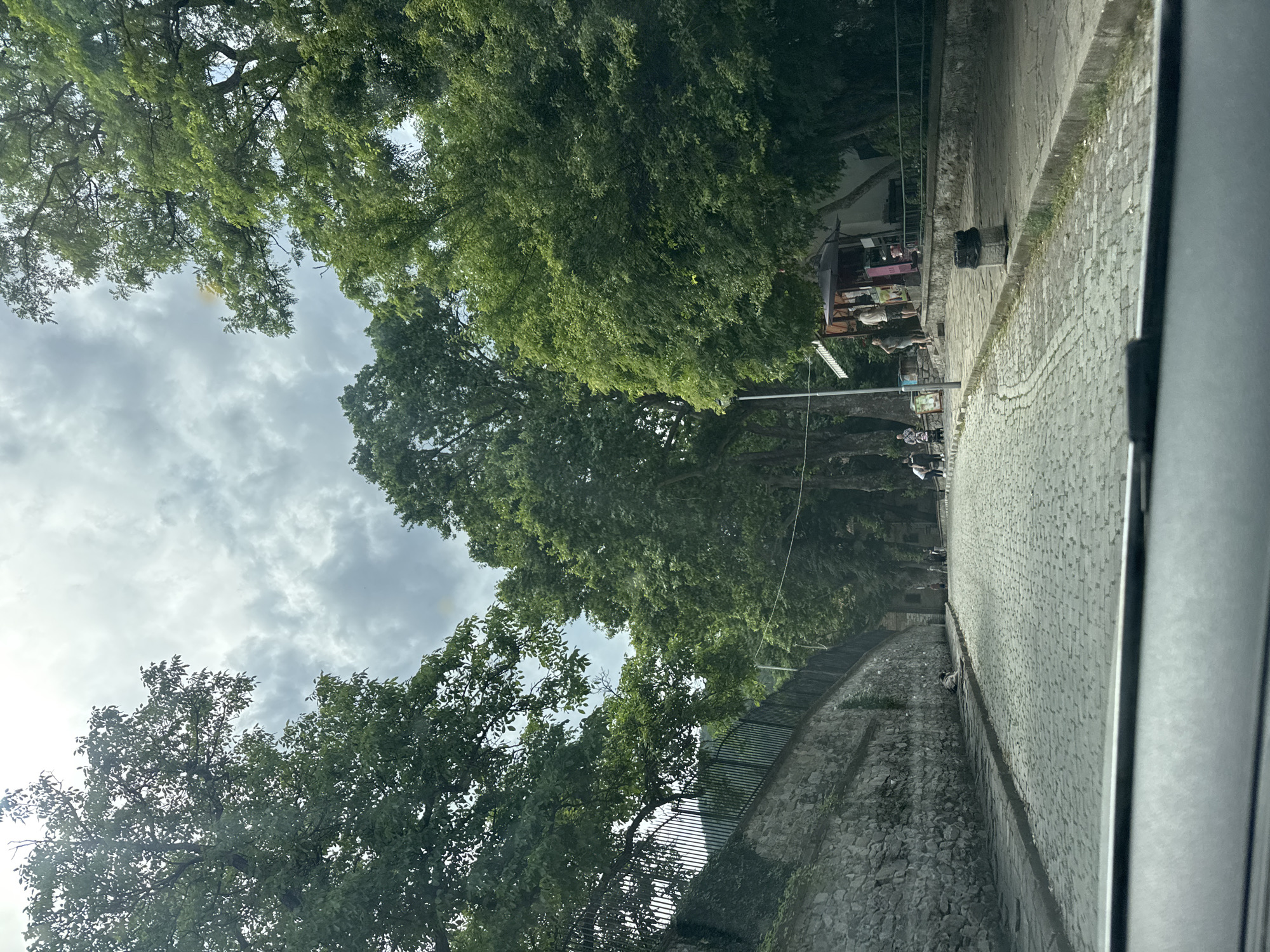 The image shows a cobbled street in Bachkovo, Bulgaria, lined with stone walls and large trees. On the right side of the street, there are a few kiosks with people standing around them. Several people are also visible walking on the sidewalk and the street. The sky is cloudy. The image is taken from a vehicle.