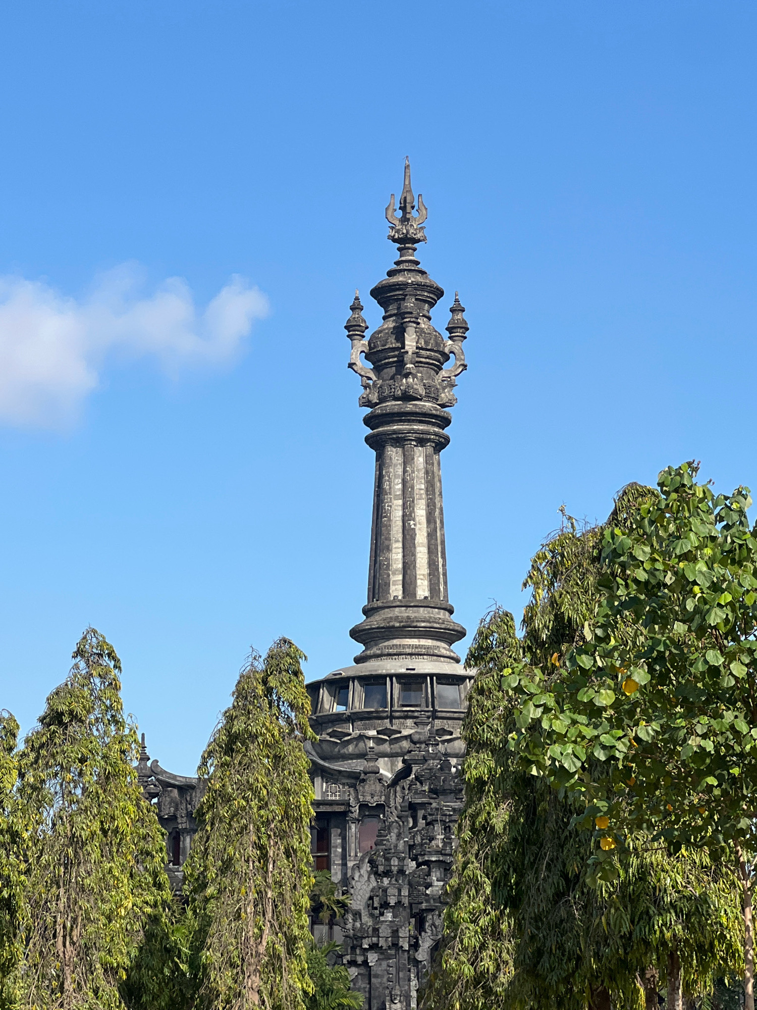 This image shows a close-up of the towering monument known as Bajra Sandhi Monument in Denpasar, Bali, Indonesia. The monument, a symbol of Balinese struggle for independence, is constructed from dark stone and features intricate carvings. The top of the monument is adorned with a distinctive spire. In the foreground, lush green trees with dense foliage partially obscure the lower sections of the monument, adding a natural element to the scene. The sky above is a clear, bright blue with a few wispy white clouds. The lighting suggests it is daytime, likely with the sun at an angle that casts shadows.