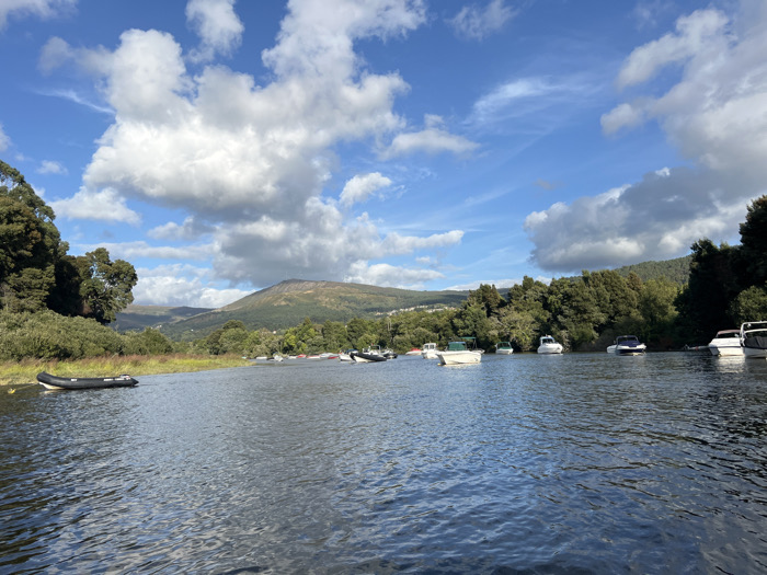 The image displays a wide view of the Minho River in Vila Nova de Cerveira, Portugal, under a blue sky with scattered cumulus clouds. The foreground is composed of dark blue water with visible surface ripples. On the left, a black inflatable boat is moored adjacent to the riverbank, which is densely covered with green trees and shrubs. Along the right bank, approximately ten white-hulled recreational boats of varying sizes are anchored or moored, extending into the mid-distance. In the background, forested green hills rise from the riverbanks, culminating in a prominent, larger mountain peak. No individuals are present. The scene is static, illuminated by daylight.
