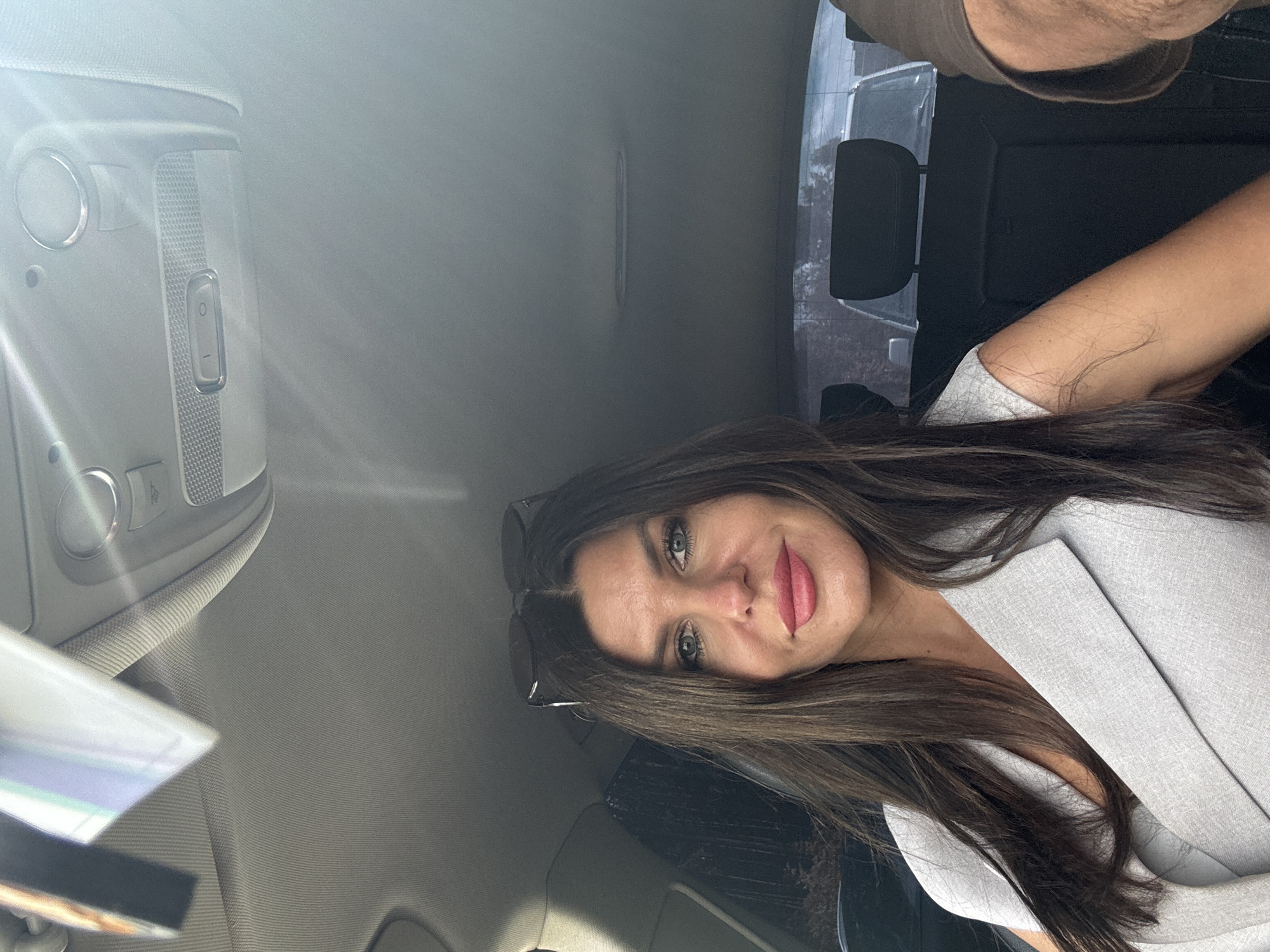 The media file shows a woman with long, dark brown hair and blue eyes taking a selfie inside a car. She is wearing a light grey blazer. Sunglasses are perched on her head. She has on bright pink lipstick and is smiling slightly. The interior of the car is visible, including the overhead console with lights and buttons. The background appears to be a slightly overcast day, with trees visible through the car's rear window. The overall mood is casual and relaxed.