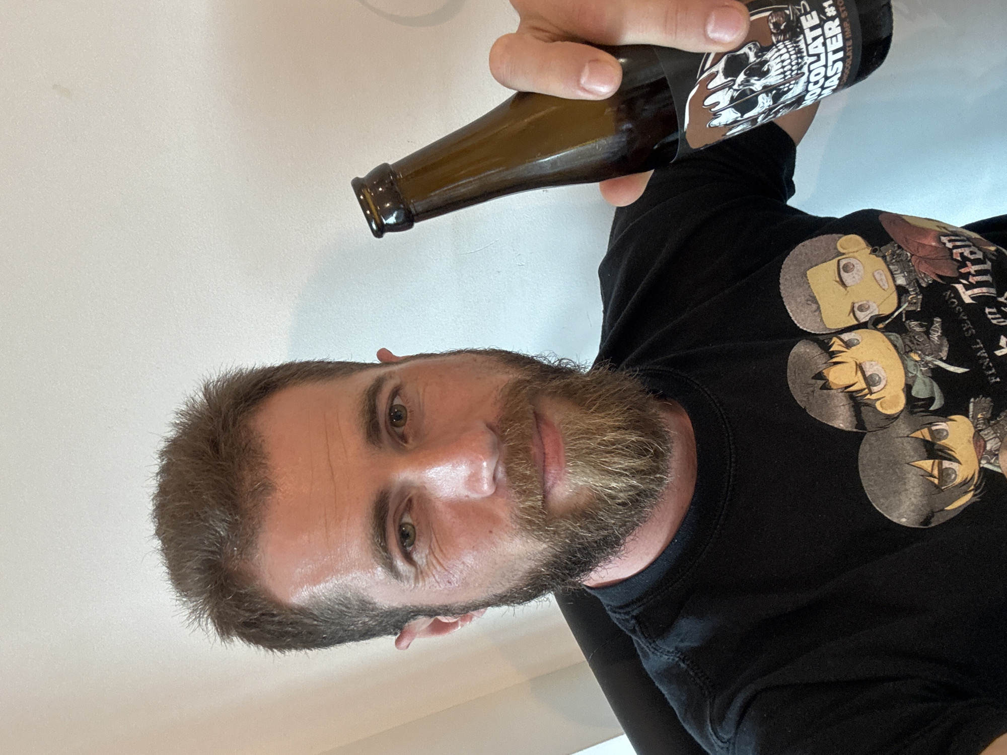 A man with brown hair and a beard is holding a bottle of "Chocolate Master #1 Chocolate Milk Stout." He is wearing a black t-shirt with "Attack on Titan" anime characters printed on it. The background is a plain white wall. There's no information in the image to determine if the photo was taken in Budva, Montenegro.