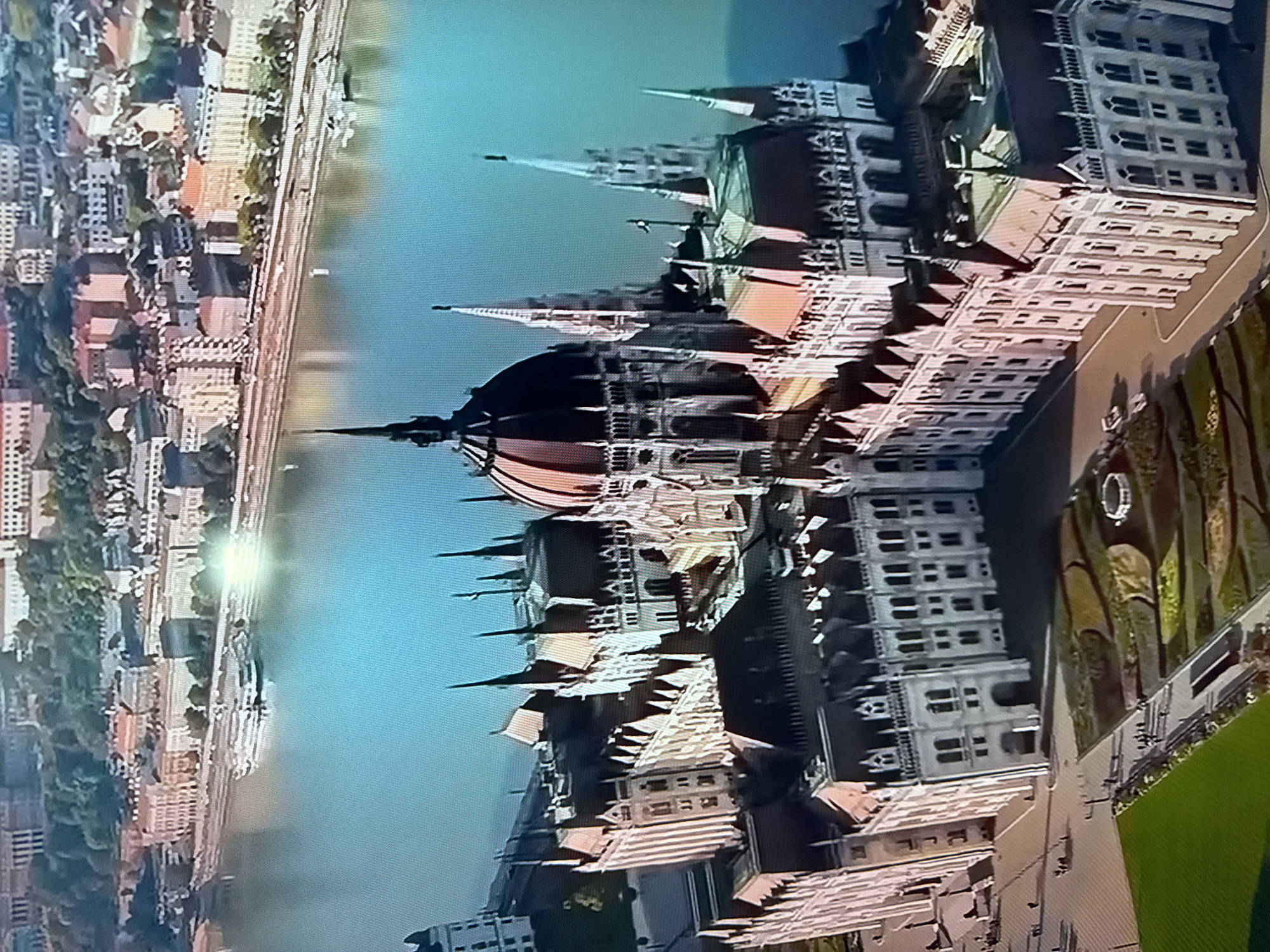This media file is an aerial, wide-angle photograph of a city with a river flowing through it. The image is oriented vertically, giving it an unusual perspective. In the foreground, the prominent subject is a large, ornate building with a domed roof and multiple spires, suggesting a landmark or significant architectural structure. The building exhibits intricate detailing and a light-colored facade.
The river, a wide expanse of blue water, is visible to the right of the building. Along the riverbank on the left side of the image, there is a dense urban area with many smaller buildings, characterized by red-tiled roofs and a mix of light and dark facades. Trees are interspersed among the buildings, indicating a green urban landscape. The overall impression is of a historical European city, likely situated on a river.
The lighting in the image suggests it was taken during the daytime, with a bright reflection on the water indicating sunlight. The sky is a clear, light blue. There are no people or specific activities clearly depicted, focusing instead on the architectural and environmental elements of the scene. The image lacks any visible text. The provided location context, "Ləcət, Azerbaijan," does not appear to align with the visual cues of the architecture, which strongly resemble buildings found in cities like Budapest, Hungary, or other Central European capitals.