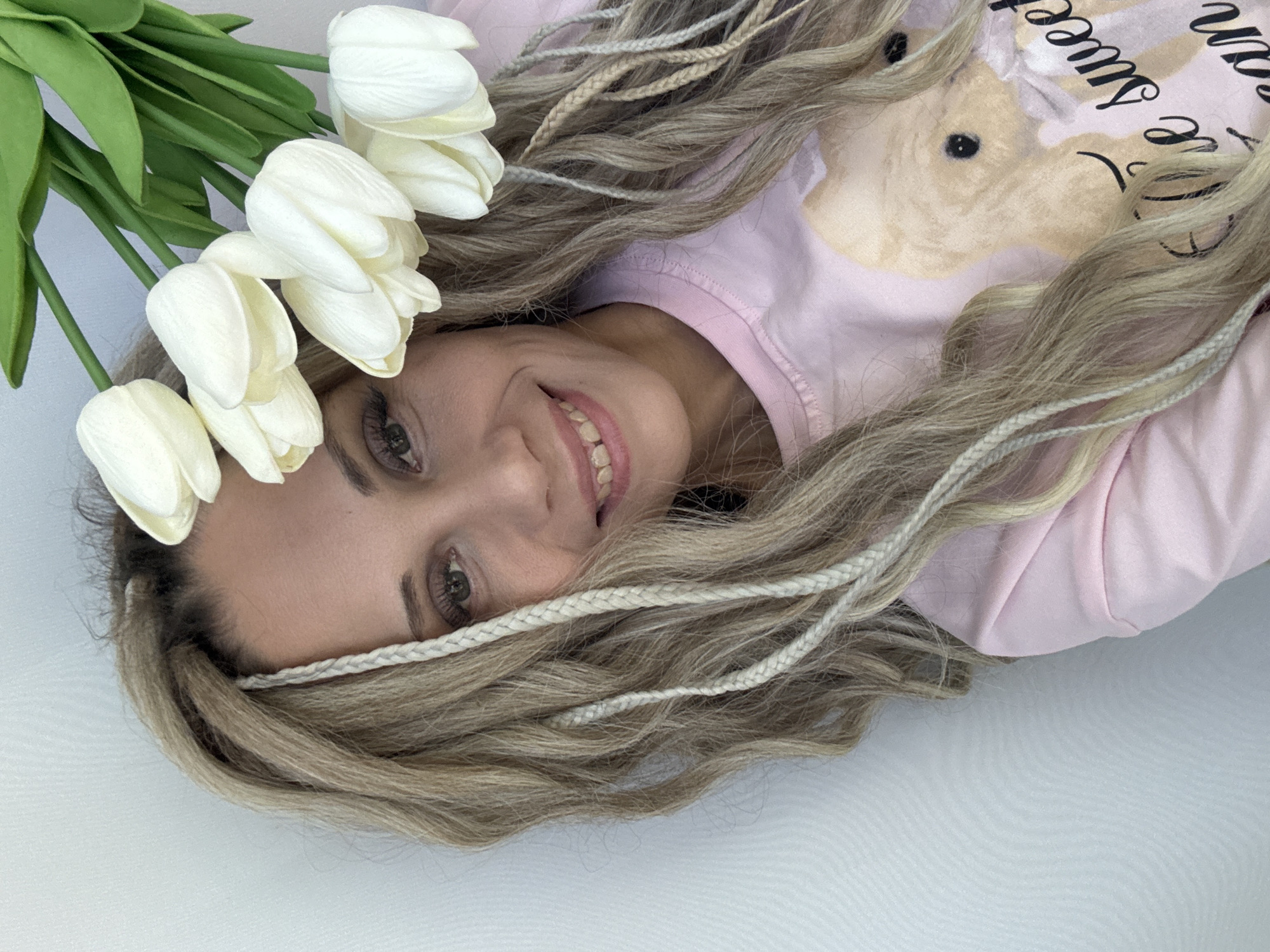 A woman lies on a white surface, her head adorned with white tulips, smiling towards the camera. She has long, braided blonde hair and wears a light pink shirt with a bunny and text on it. The image is close-up, focusing on the woman's face and upper body. The background is plain and does not indicate any specific location within the city of Unknown, Unknown.