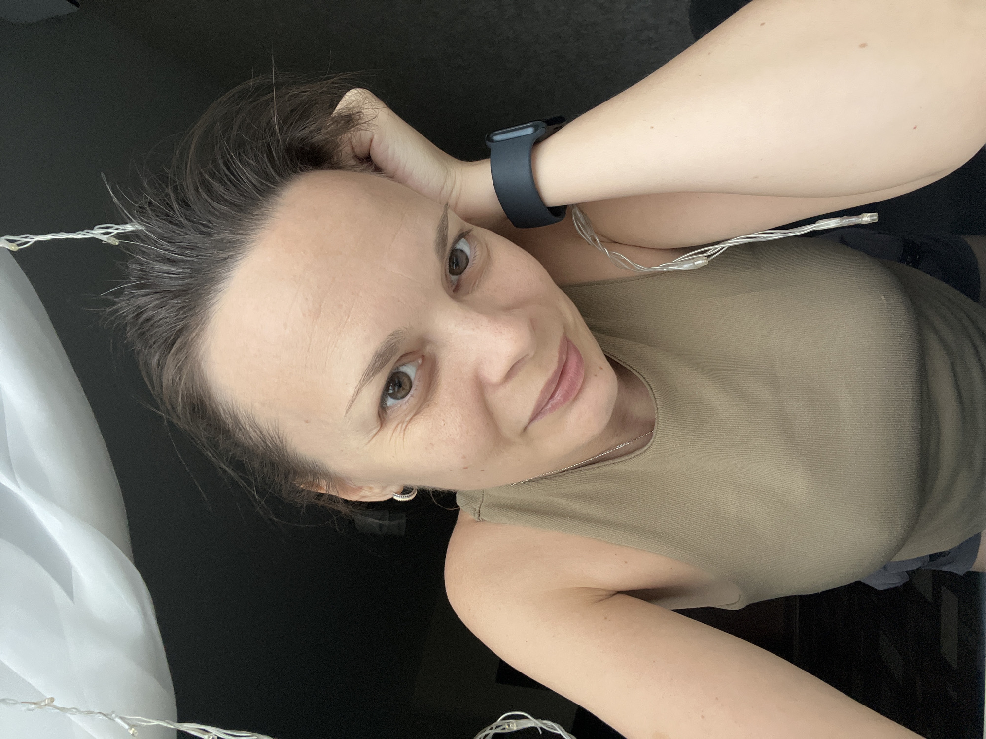 A medium shot selfie captures a woman resting her head on her hand, looking towards the camera. She has light brown hair, light brown eyes, and fair skin with freckles. She is wearing a tan tank top and a dark smartwatch on her left wrist. String lights are visible around her. The background is dark, with a glimpse of a white curtain in the upper left corner. The scene does not provide any specific indicators that would allow for the identification of the location as Tolyatti, Russia.