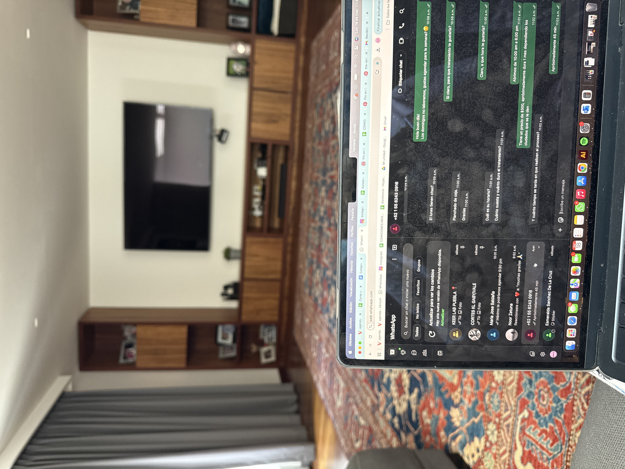 Here's a detailed description of the image:
**Scene:** The image shows a room interior with a focus on a laptop screen displaying a WhatsApp chat. The background includes elements of a living space:
* A large built-in wooden shelving unit with a television mounted in the center.
* A gray curtain visible in the lower left corner, suggesting a window.
* A portion of a colorful rug or carpet in the foreground.
**Laptop Screen (WhatsApp Chat):**
* The laptop is running the WhatsApp web application in Chrome.
* The screen shows a chat with "+52 1 55 6243 0916".
* The conversation includes inquiries about scheduling an appointment, specifically for "planchado de ceja" (eyebrow shaping), pricing, and duration of the treatment.
* The chat also mentions that the business is closed on Sundays, and hours of operation from 10:00 am to 8:00 pm.
* The messages indicate a price of $300 and a treatment duration of approximately one month, with the actual duration dependent on the clients.
* The conversation takes place in real-time, as the chat messages have timestamps around 10:59 a.m. to 11:03 a.m.
**Additional Details:**
* The WhatsApp interface also shows other contacts, including "KEER LAB PUEBLA," "CORTES KL GABY/VALE," "Maria Jose Saldaña," and "Noor Zetuna."
* The Chrome browser has several tabs open, including agenda pages, email, and other web applications.
* The laptop's dock shows a variety of applications, including Adobe Illustrator, Spotify, and other common software.
**Location (Implied):**
* Based on the WhatsApp contact "+52 1 55 6243 0916," the location is likely Mexico, as +52 is the country code.
* One of the WhatsApp contact names is "KEER LAB PUEBLA," suggesting the location may be in or near Puebla, Mexico.
* There is no mention of San Bernardino Tlaxcalancingo, Mexico.