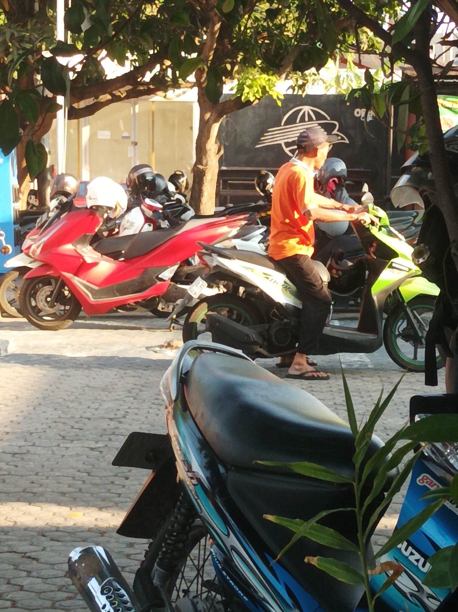 A street scene in Nganjuk, Indonesia, features a variety of parked and ridden motorcycles under dappled sunlight filtering through tree branches. In the foreground, a person wearing an orange shirt and dark pants is seated on a green and white scooter, with their right hand on the handlebars. A red scooter is parked to the left of the green one. Behind these scooters, several other motorcycles and helmets are visible. The ground is paved with interlocking bricks. In the background, a building with glass panels and a dark wall with a white graphic are visible. The overall impression is of a busy urban environment where motorcycles are a primary mode of transportation.