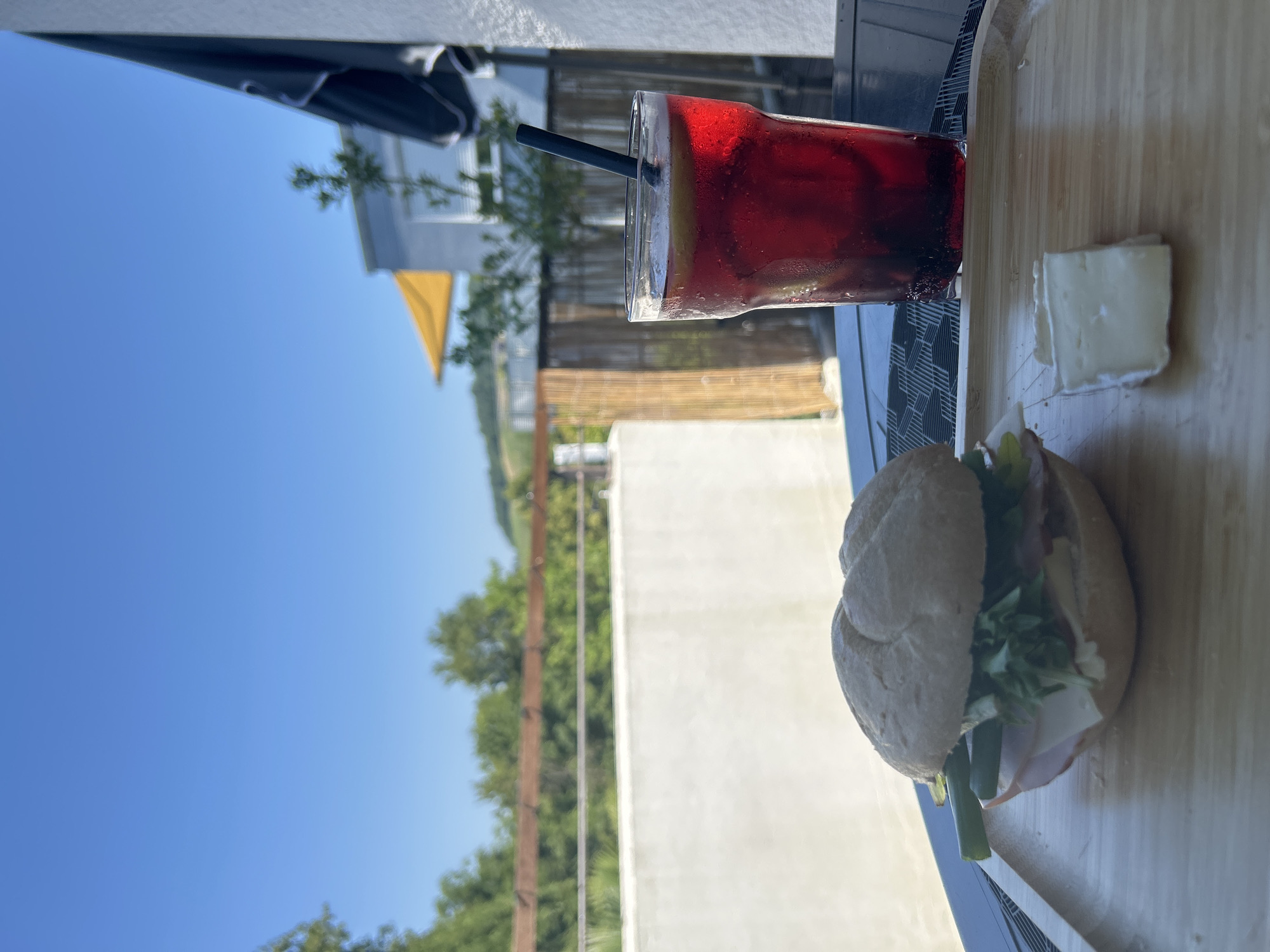 A close-up, eye-level shot shows a sandwich and a glass of red beverage on a wooden tray. The sandwich, on the left, is a small bun with lettuce, ham, cheese, pickles, and green beans. Next to it, on the right, is a tall glass filled with a dark red beverage, ice, lemon slices, and a black straw. A small piece of white cheese is also on the tray to the right of the sandwich. In the background, a white wall and a wooden fence are visible, with trees and a distant hillside under a clear blue sky. The scene appears to be outdoors, possibly on a balcony or patio.