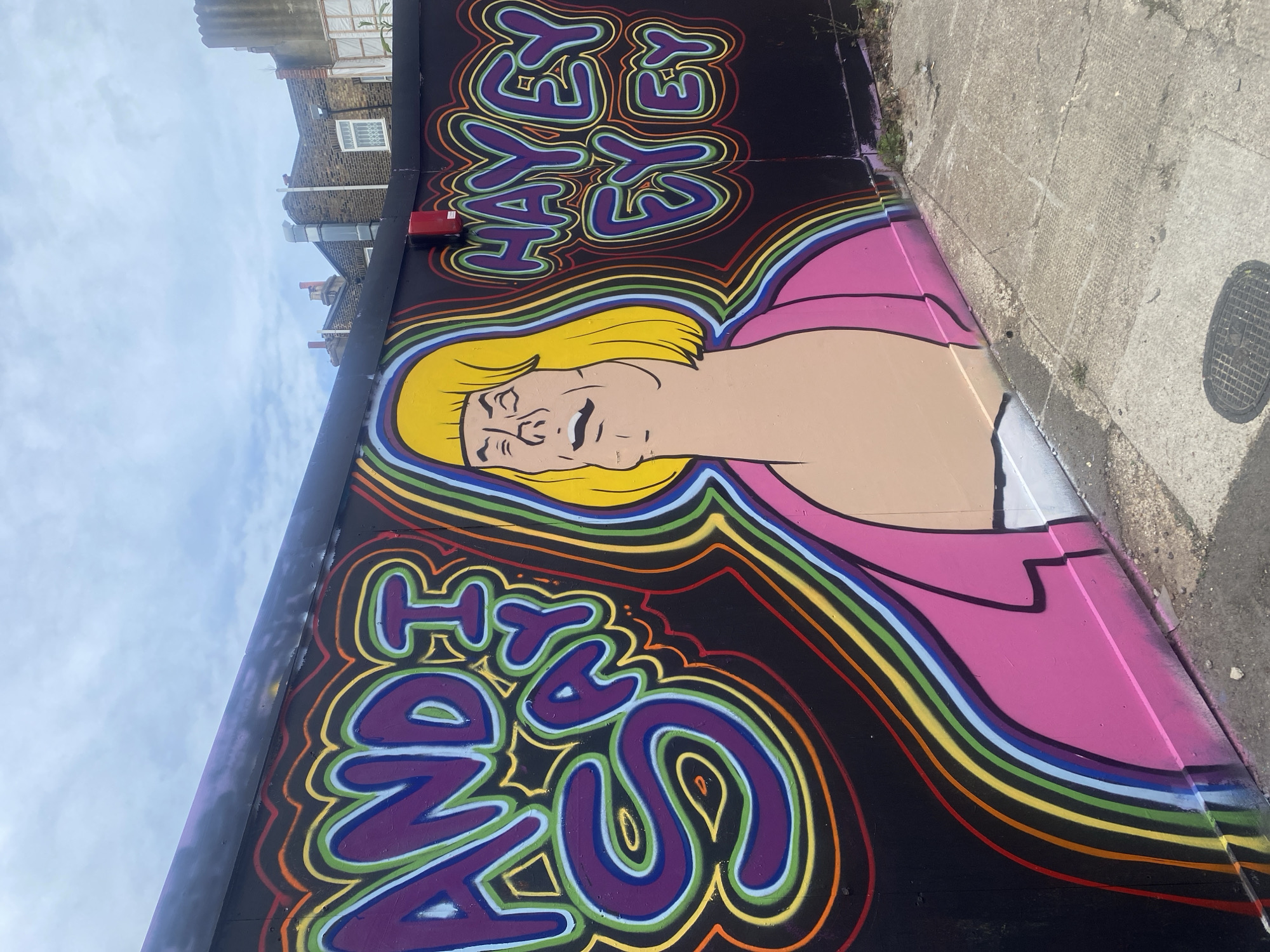 A section of street art in London, United Kingdom, depicting a portrait of a man with blonde hair and a pink garment. The text "And I say hey yeah yeah yeah hey yeah yeah" is rendered in a psychedelic font around the portrait. The artwork is painted on a wall adjacent to a sidewalk. Buildings and sky are visible in the background.